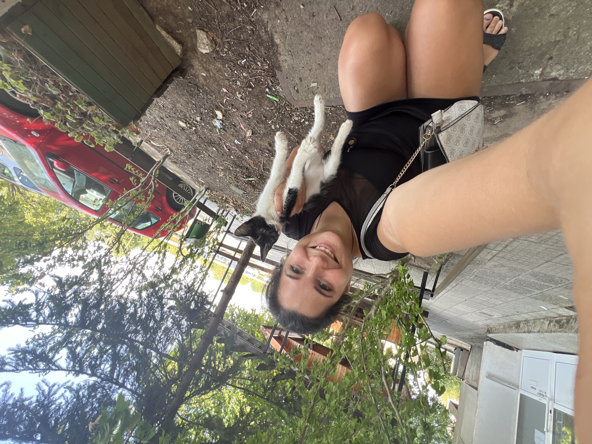 A woman is taking a selfie while holding a black and white kitten outdoors. She is wearing a black dress and sandals, and carrying a small purse. The kitten is held in her arms, looking towards the camera. In the background, there is a red car, trees, and a building. The setting appears to be a street or walkway in the city of Vratsa, Bulgaria.
