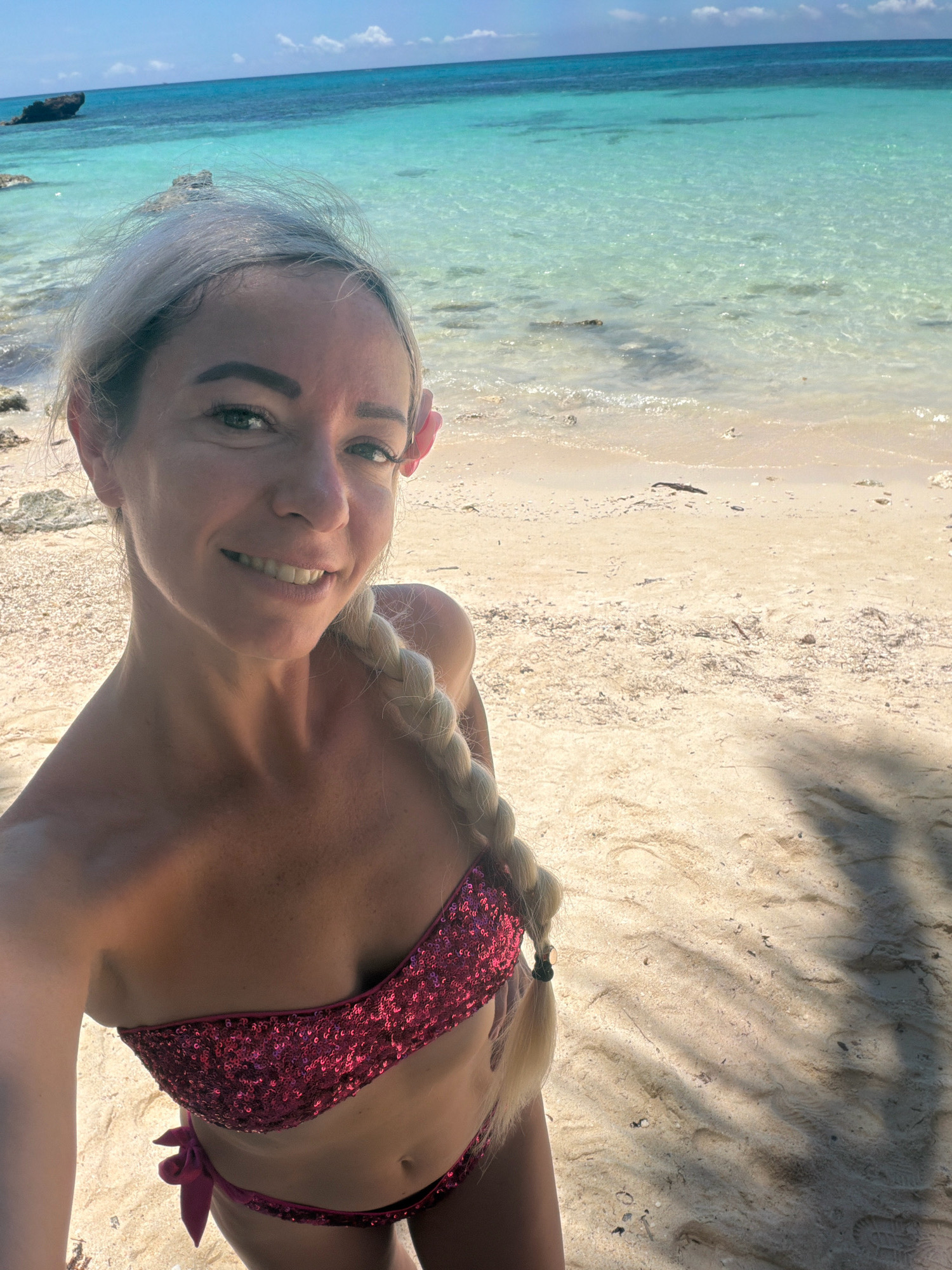 A woman with blonde hair, styled in a braid and adorned with a pink flower behind her ear, takes a selfie on a sandy beach. She is wearing a sparkling red bikini. The ocean, with its clear turquoise water, stretches out behind her, meeting a bright blue sky with scattered clouds. The beach is light-colored sand with small shells and debris scattered across it. Shadows of palm fronds are visible on the sand, suggesting the presence of nearby trees. The image appears to be taken in a tropical location.