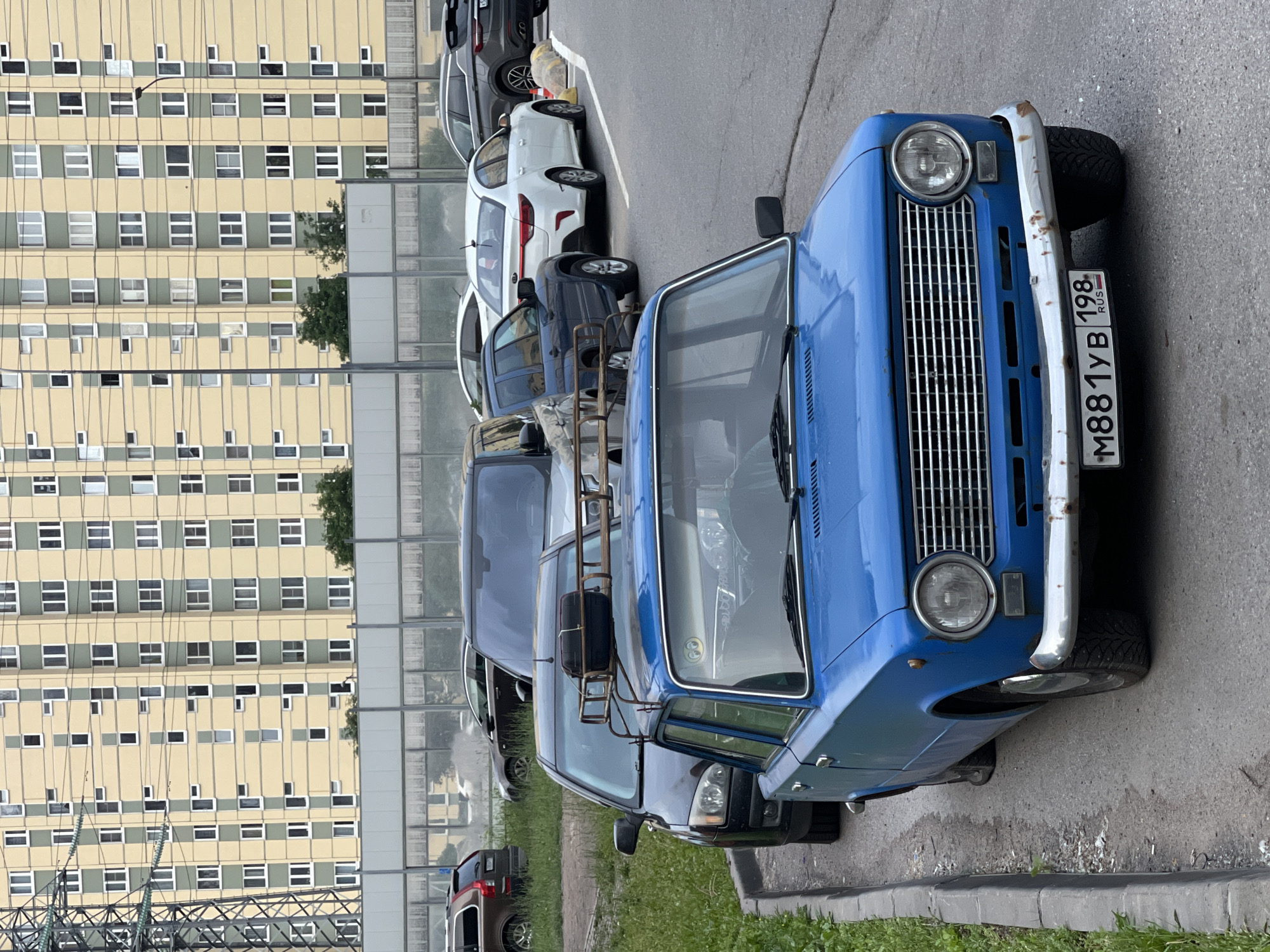 A blue VAZ-2106 car, license plate M881YB 198, is parked on the right side of a road in an urban setting. The car has a roof rack with a black suitcase on it. The car is positioned in the foreground, partially obscuring other vehicles parked behind it, including a dark-colored Ford Focus and a dark blue SUV. In the background, a large, light-yellow apartment building with green window frames dominates the scene. A power line tower is visible on the far left. The ground is asphalt, with a grass verge and a concrete curb on the left side. The sky is overcast.