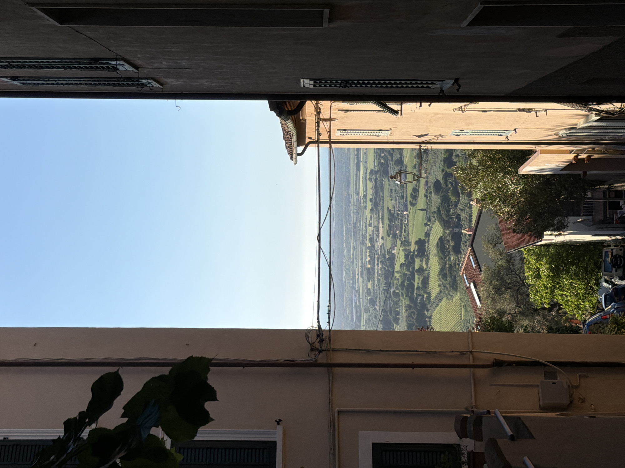 The image depicts a view framed by two buildings, looking out over a rural landscape. The buildings are light colored with dark green shutters. Overhead wiring and pipes are visible along the lower building. The landscape is dominated by green fields and wooded areas, under a clear blue sky. A building with a red tile roof is visible amidst the greenery. A road winds through the landscape. Parked cars are partially visible on the right side of the frame. The image is likely taken in Castagneto Carducci, Italy, based on the typical architecture of the buildings and the landscape.