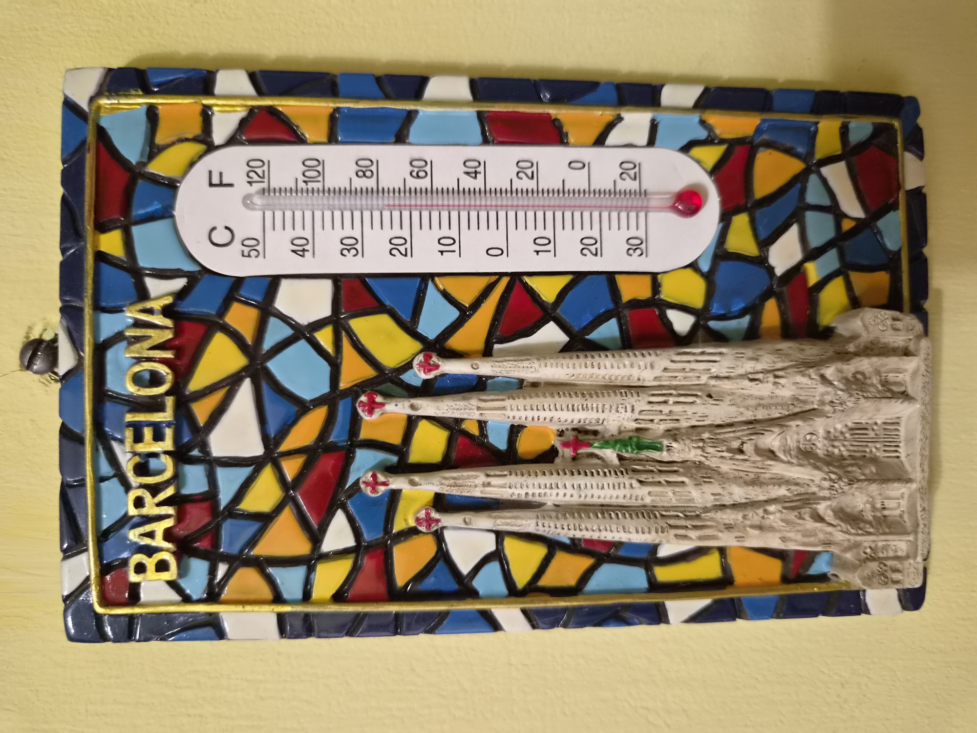 The image displays a decorative thermometer with a stained-glass background and a miniature representation of the Sagrada Familia basilica. The thermometer is attached to a yellow wall and features markings for both Celsius and Fahrenheit. The red liquid in the thermometer indicates the temperature. The text "BARCELONA" is prominently displayed in gold lettering above the stained-glass mosaic. A small insect is visible at the top of the frame. The scene is indoors.
