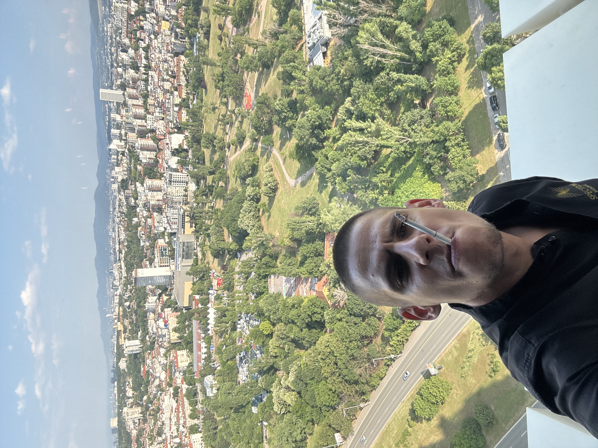 A man is taking a selfie on a balcony, holding a cigarette in his mouth. He is wearing a black collared shirt with a logo on the left side. In the background, there is a panoramic view of Sofia, Bulgaria. The city features a dense urban landscape with numerous buildings of varying heights, interspersed with green spaces, parks, and trees. A highway runs through the foreground, with cars visible on the road. The sky is blue with scattered clouds, and mountains are visible in the distance. The lighting suggests it is daytime, possibly late afternoon.