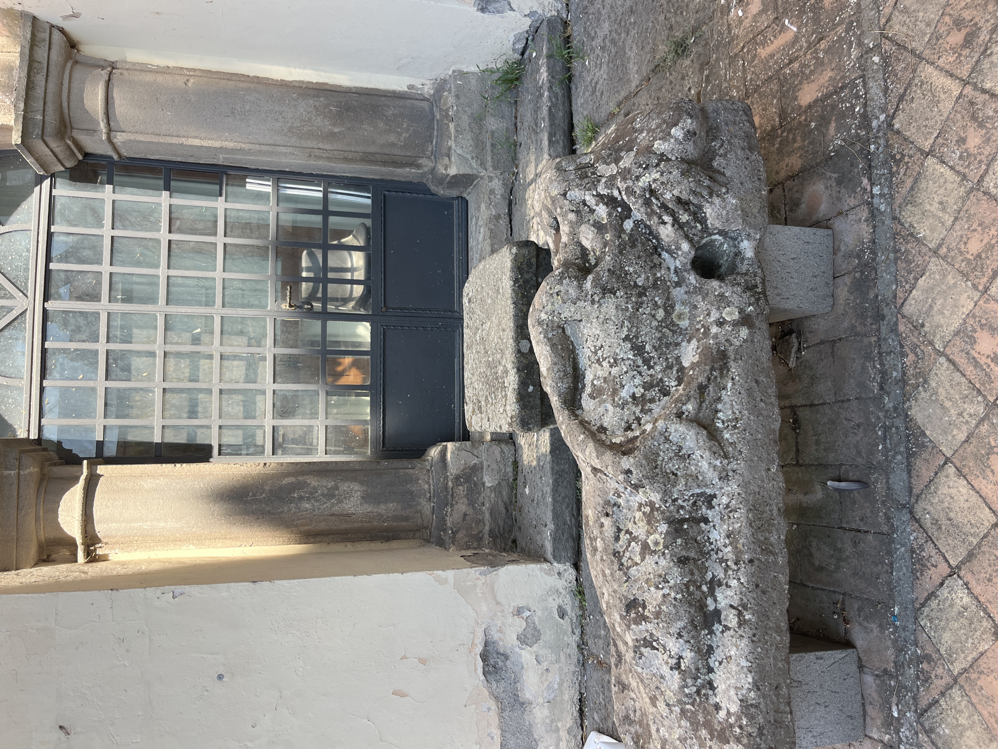 In Viterbo, Italy, in front of a building with a weathered stucco facade and a large, multi-paned metal door, rests a weathered stone sarcophagus. The sarcophagus, carved to depict a reclining figure, is covered in moss and lichen, and has a noticeable hole on its right side. It is supported by two small stone blocks and sits on a brick patio. Behind the sarcophagus, a stone step leads up to the aforementioned door. The scene is bathed in natural light, casting shadows from the architectural elements.