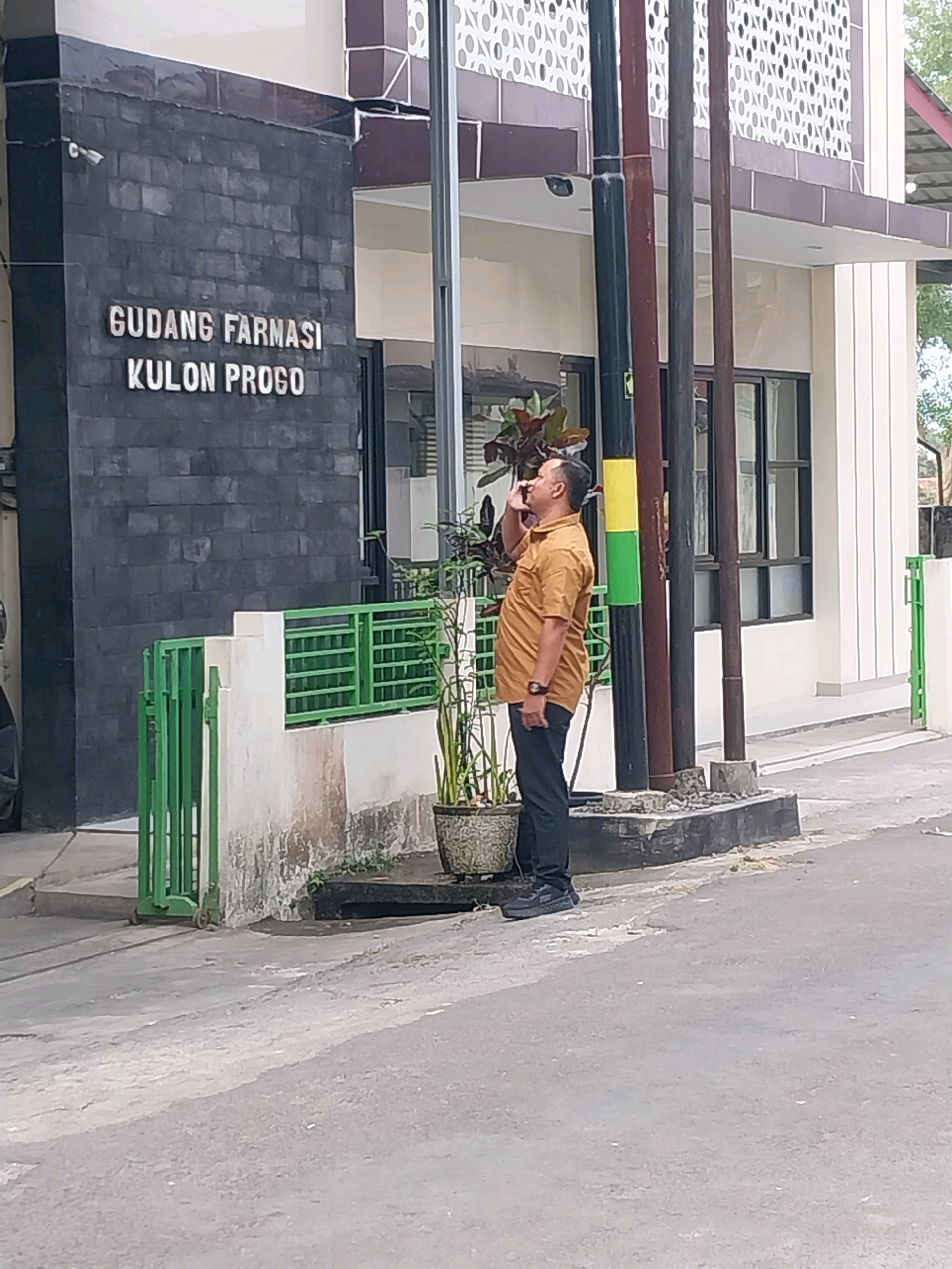 The image captures a full-body view of a man standing on a sidewalk in front of a building. The man is wearing a short-sleeved, tan-colored shirt and dark pants. He appears to be smoking and looking upwards and to his left. The building behind him has a dark brick facade with white lettering that reads "GUDANG FARMASI KULON PROGO". To the left of the entrance, a security camera is mounted on the brick wall. In front of the building, there is a green metal fence and some potted plants. The man is standing near a dark pole with a yellow and green stripe. The scene is located in Wates, Indonesia, as indicated by the building's signage. The ground is paved, and there is a street in the foreground.
