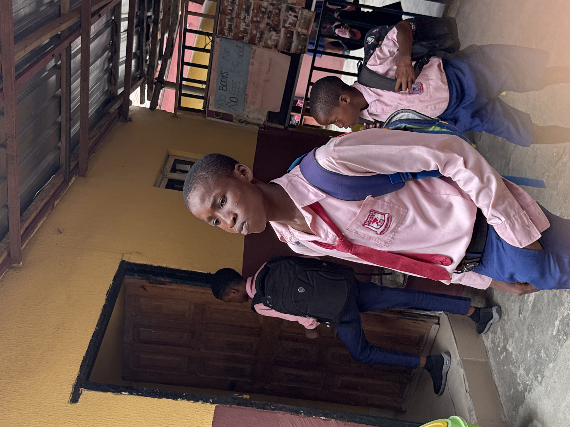 Three young students in matching pink button-down shirts, blue trousers, black shoes, and backpacks are visible in an outdoor school setting, which may be located in Ado, Nigeria. The foreground student is looking forward, his shirt is untucked, and his red tie is loosely worn. He wears a blue backpack. Another student walks behind the main subject wearing a black backpack. Behind them, a third student is partially visible. A sign is posted near the second student, reading, "NO BOOKS, NO FEES, SCHOOL." They are near a building, entering through a wooden door on a yellow wall. The building's roof appears to be made of corrugated metal.