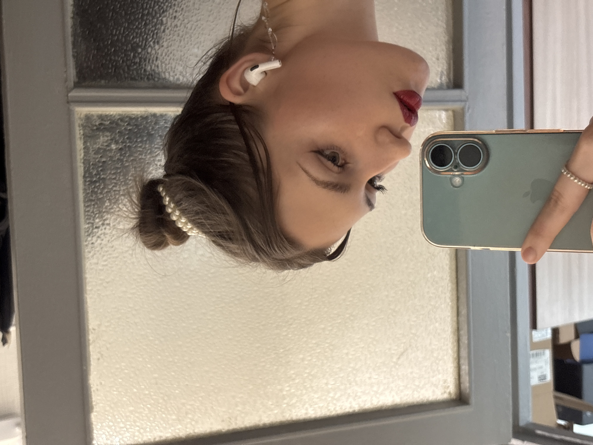 The image captures a young woman in profile, positioned in an indoor setting within Saint Petersburg, Russia, as she takes a mirror selfie. Her dark hair is styled into a neat bun, adorned with a decorative string of white, pearl-like beads. She wears bold red lipstick and prominent winged eyeliner. In her visible right ear, a white Apple AirPod Pro is inserted, from which a clear, elongated decorative element resembling an earring dangles. Her left hand, featuring a silver-colored beaded ring on the ring finger, holds a light green smartphone with visible dual camera lenses, reflecting its back towards the viewer, indicating she is photographing her own reflection. The background, also a reflection, reveals a textured, possibly frosted glass panel set within a light grey door or window frame, with hints of an interior space featuring stacked items or shelves faintly visible on the far left.