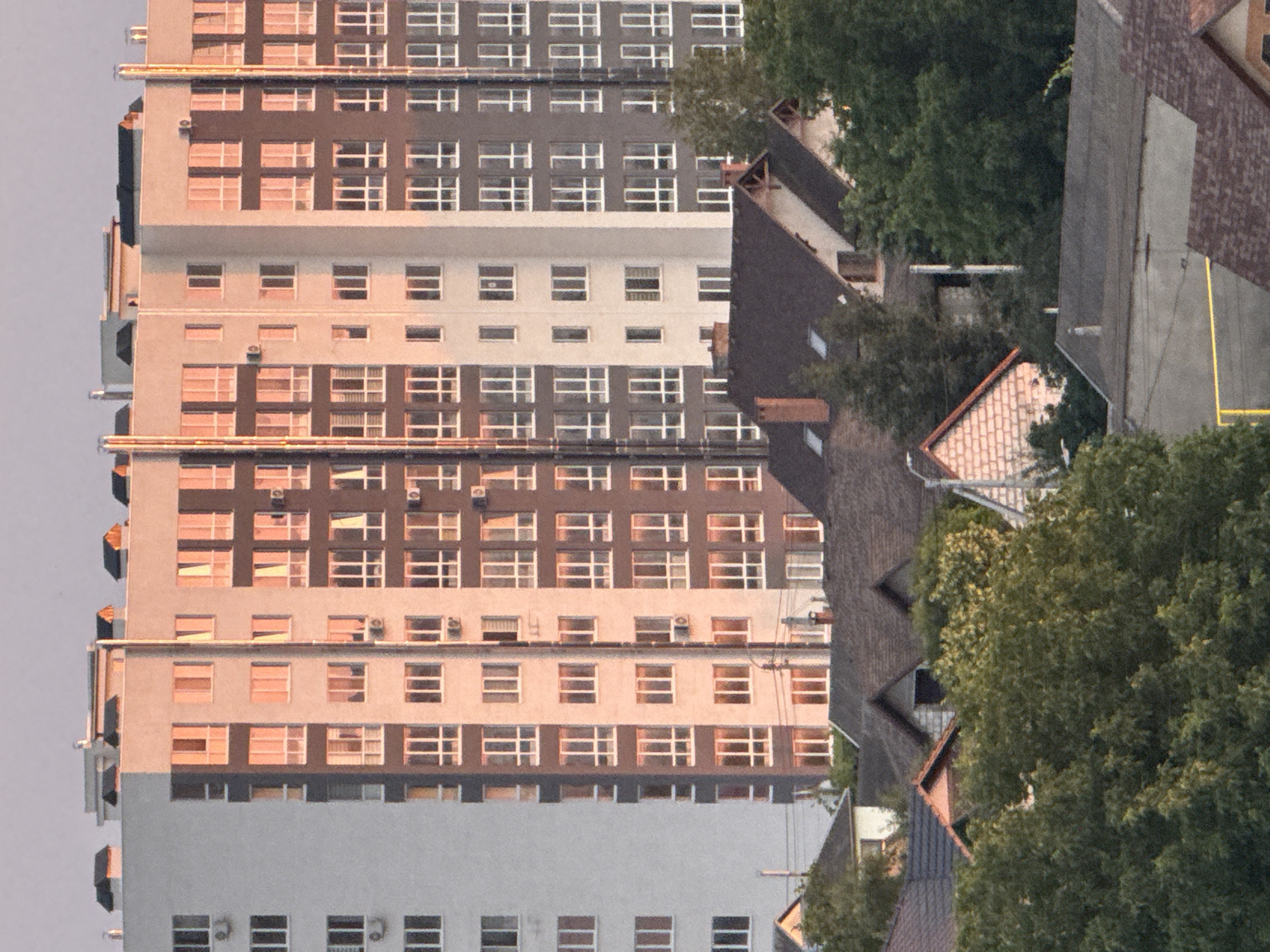 The image depicts a residential building in Cahul, Moldova. The building is tall and modern, with a light-colored facade that is partially illuminated by the warm glow of sunset. The building features numerous windows, many of which are reflecting the sky and surrounding environment. Below the tall building, there are several smaller houses with dark roofs, partially obscured by lush green trees. The scene appears to be a typical urban or suburban landscape, with a focus on residential architecture. There are no people visible in the image, and the environment suggests a calm and peaceful atmosphere.
