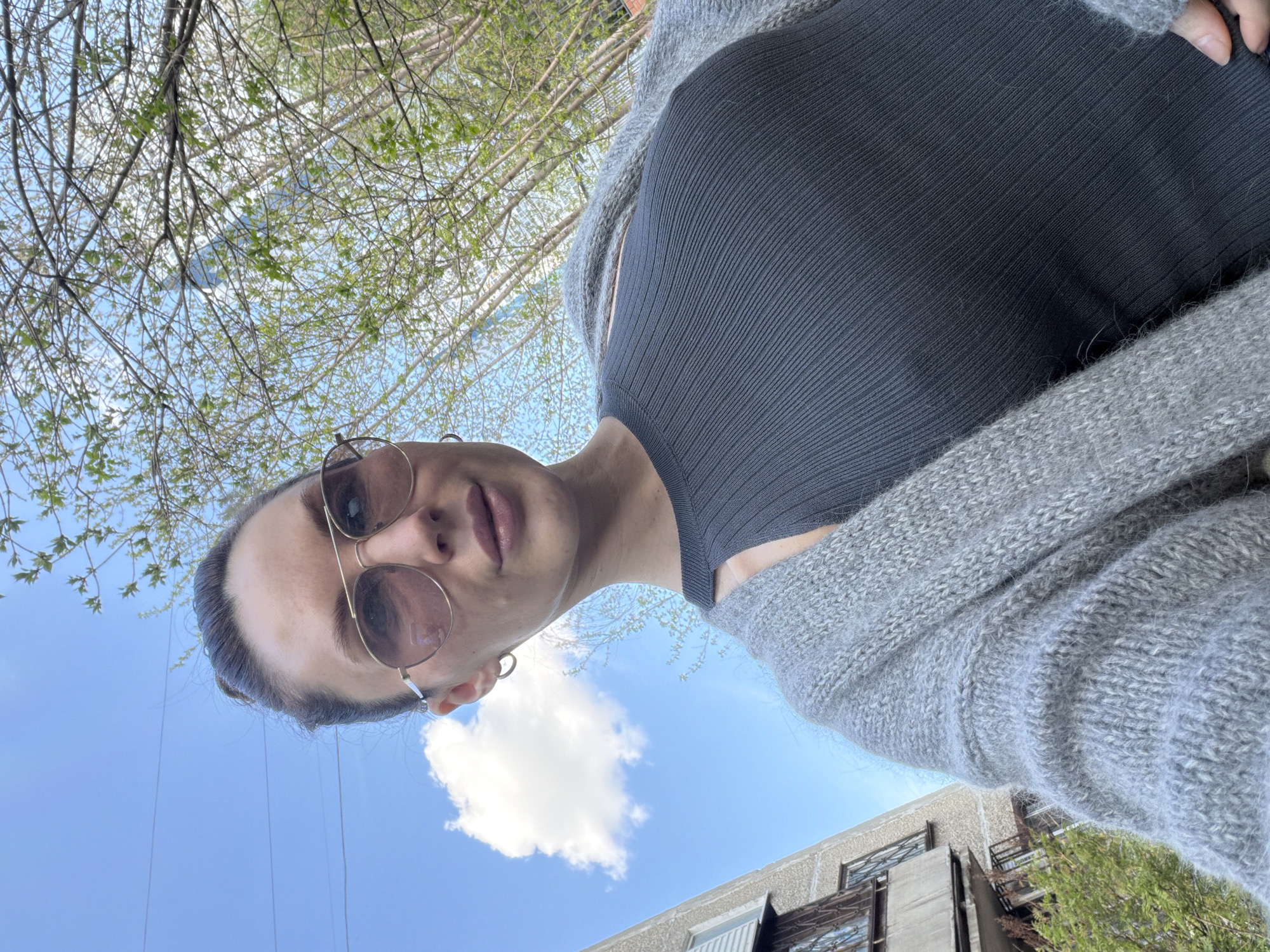 A woman is looking down at the camera, wearing sunglasses, a gray sweater, and a dark gray top. The background includes a building, a tree with new green leaves, a blue sky with a white cloud, and thin white power lines. The photograph appears to have been taken outdoors, looking upwards. It is not possible to determine the location of Perm, Russia from the image provided.