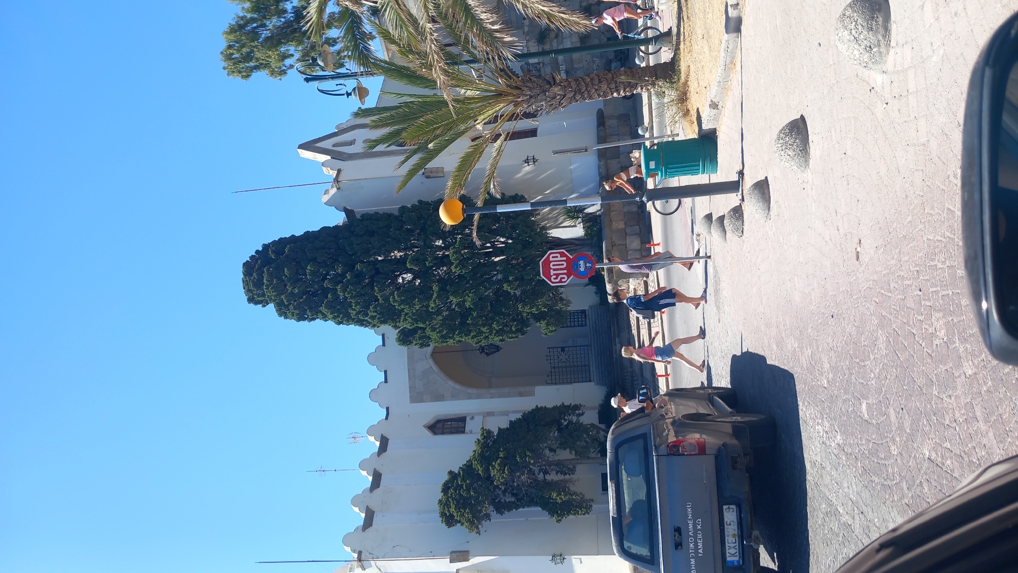 A daylight, eye-level, medium shot shows a busy intersection in the city of Kos, Greece, on a clear sunny day. The foreground is dominated by a gray pickup truck with a license plate reading "KXE 75 9." Behind the truck, a group of people are crossing the street. A woman in a pink top and denim shorts walks across, followed by two men in casual wear. Further to the right, a woman on a bicycle passes a green trash can. A palm tree and a tall cypress tree stand near a white building with a prominent arched entrance. A red "STOP" sign is visible on a pole, along with a yellow street lamp. The street is paved with cobblestones. The building in the background appears to be a public building, possibly administrative or historical, given its architecture.