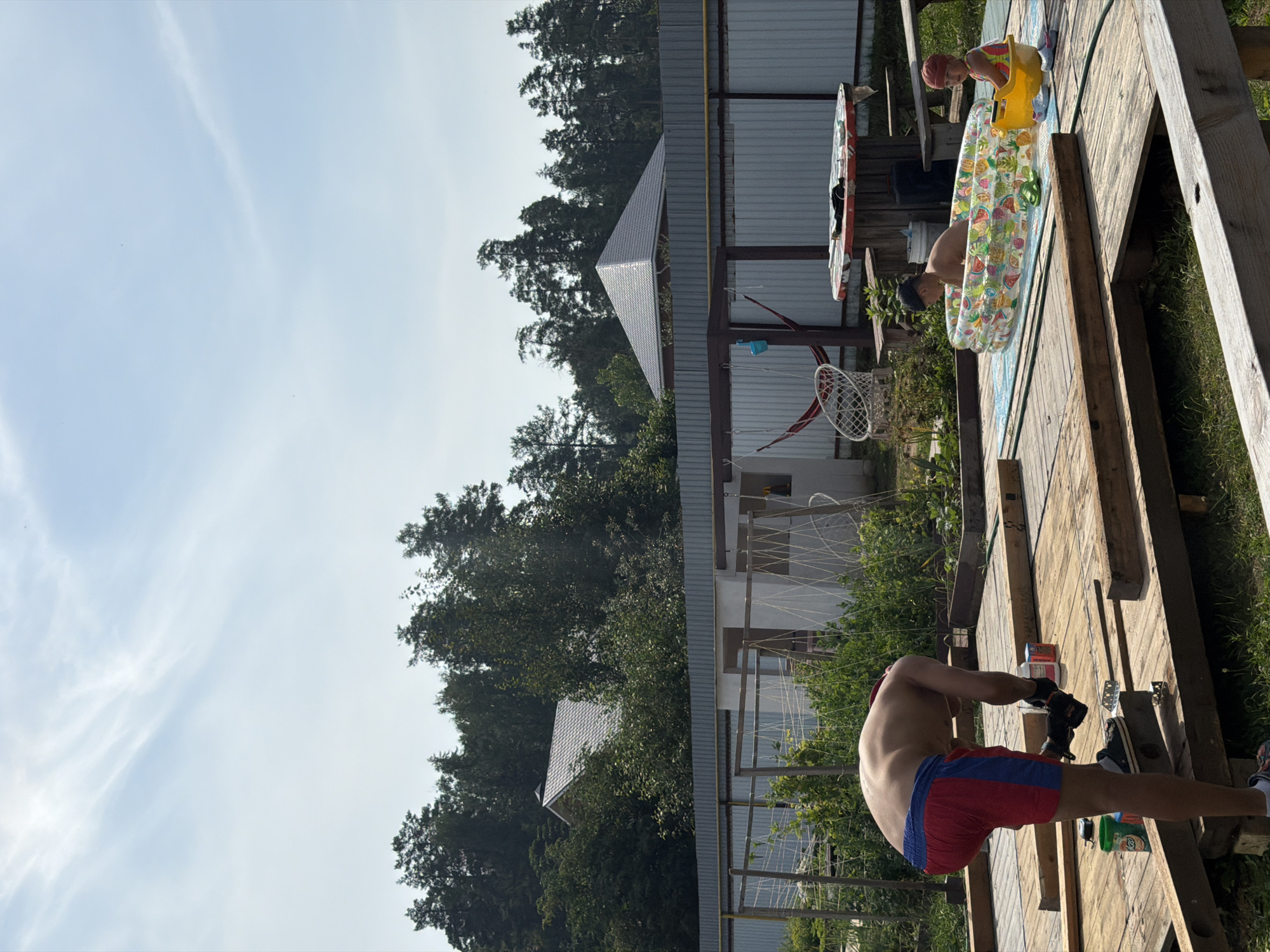 The image depicts a backyard scene in Chaikovsky, Russia, on a sunny day. A young man, shirtless and wearing red and blue shorts, is engaged in construction work on a wooden deck, holding a power tool. In the background, two children are playing in an inflatable pool. One child is submerged in the water, while the other sits in a yellow toy boat within the pool. A picnic table with chairs is visible nearby, along with a hammock suspended from a wooden frame. The area is surrounded by a metal fence and lush greenery, with trees and a house visible in the distance.