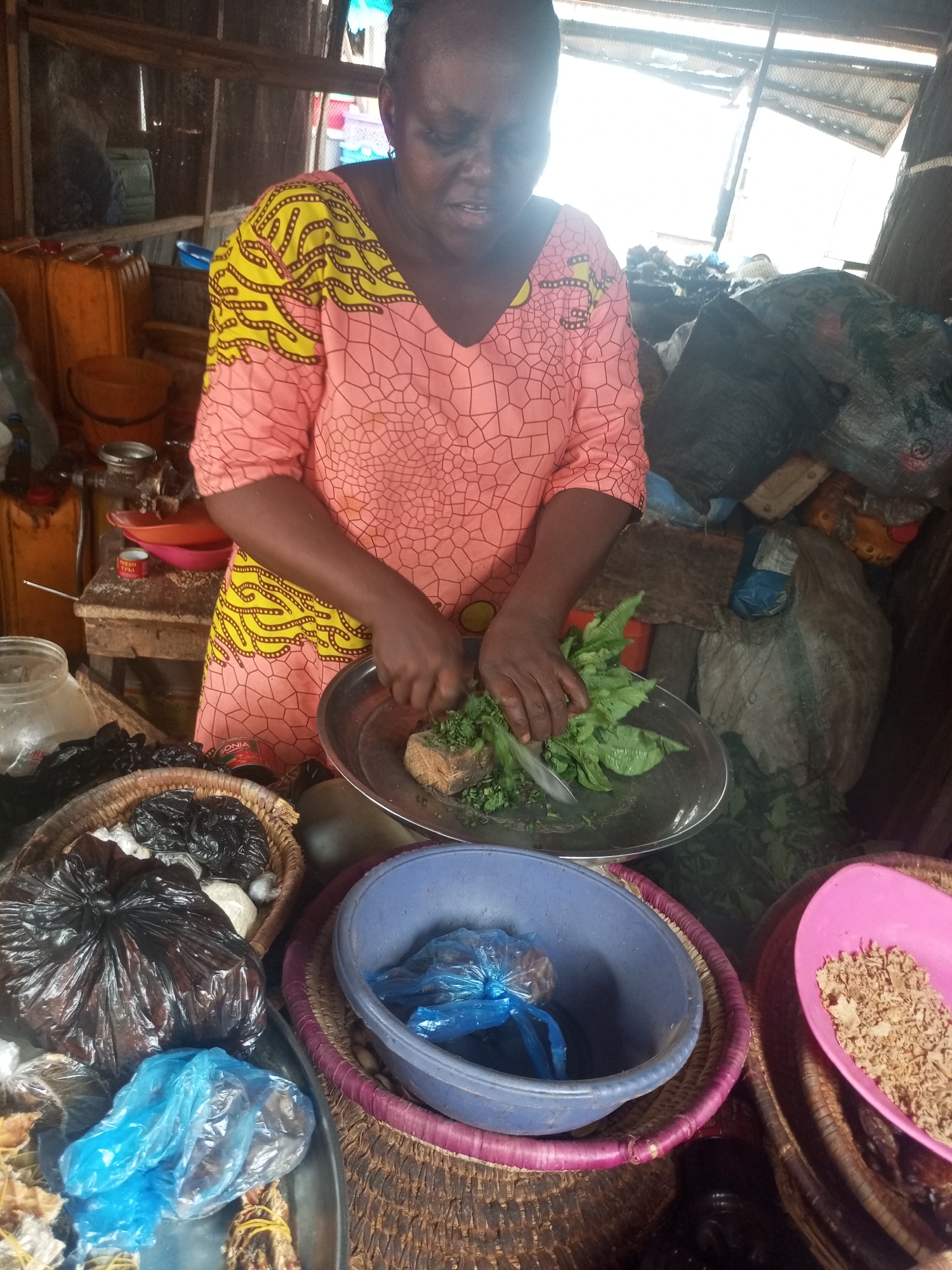 In an indoor market scene in Sabon Tasha, Nigeria, a middle-aged Black woman wearing a patterned pink and yellow dress is meticulously chopping green vegetables with a knife on a silver plate. Around her are several woven baskets and plastic bowls containing various goods for sale, including dried foods and items wrapped in plastic bags. The market stall is cluttered with items stacked behind her, including yellow containers and bundles of unidentified goods. The environment is dimly lit, suggesting a basic market setup.