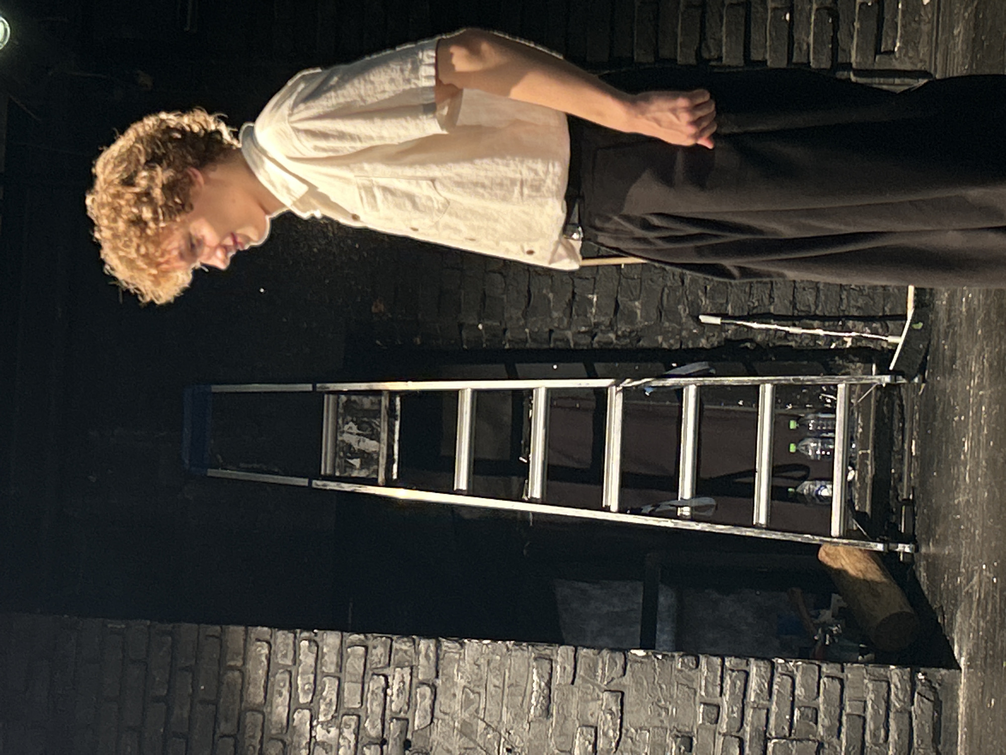 A young person with curly blond hair, wearing a loose-fitting cream-colored shirt and dark pants, is seen from the side, leaning over and looking downwards. Their left arm is bent, and their right arm is down by their side. They appear to be on a stage, suggested by the brick-like backdrop and the presence of a metal ladder. The ladder is set up in front of the brick wall and has several rungs. On one of the upper rungs of the ladder, there are several plastic water bottles. Below the ladder, a thick piece of wood, possibly a log, is visible. The lighting suggests an indoor setting, possibly a theater or performance space, with a spotlight illuminating the person and parts of the ladder. The overall mood is somewhat somber, given the dim lighting and the person's downward gaze. There is no visible text in the image.
