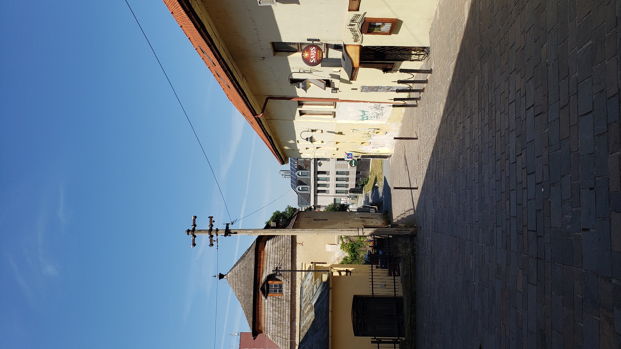 The image depicts a street scene in Prešov, Slovakia. The perspective is from a slightly elevated position looking down a cobblestone street. To the left, an older, light-yellow building with a distinctive multi-faceted wooden roof dominates the foreground. A dark metal fence is in front of it, and a dark wooden door is visible. To the right, a lighter yellow building with a red-tiled roof angles into the frame. A sign for "SARIS" is visible on this building, along with another sign that reads "RESTAURANT & PIZZA". The street is lined with black bollards and chains in some areas. In the background, other buildings can be seen, including one with solar panels on its roof. The sky is clear and blue with a few wispy clouds. A utility pole with wires is prominent in the center of the image. The scene appears quiet and unpopulated.