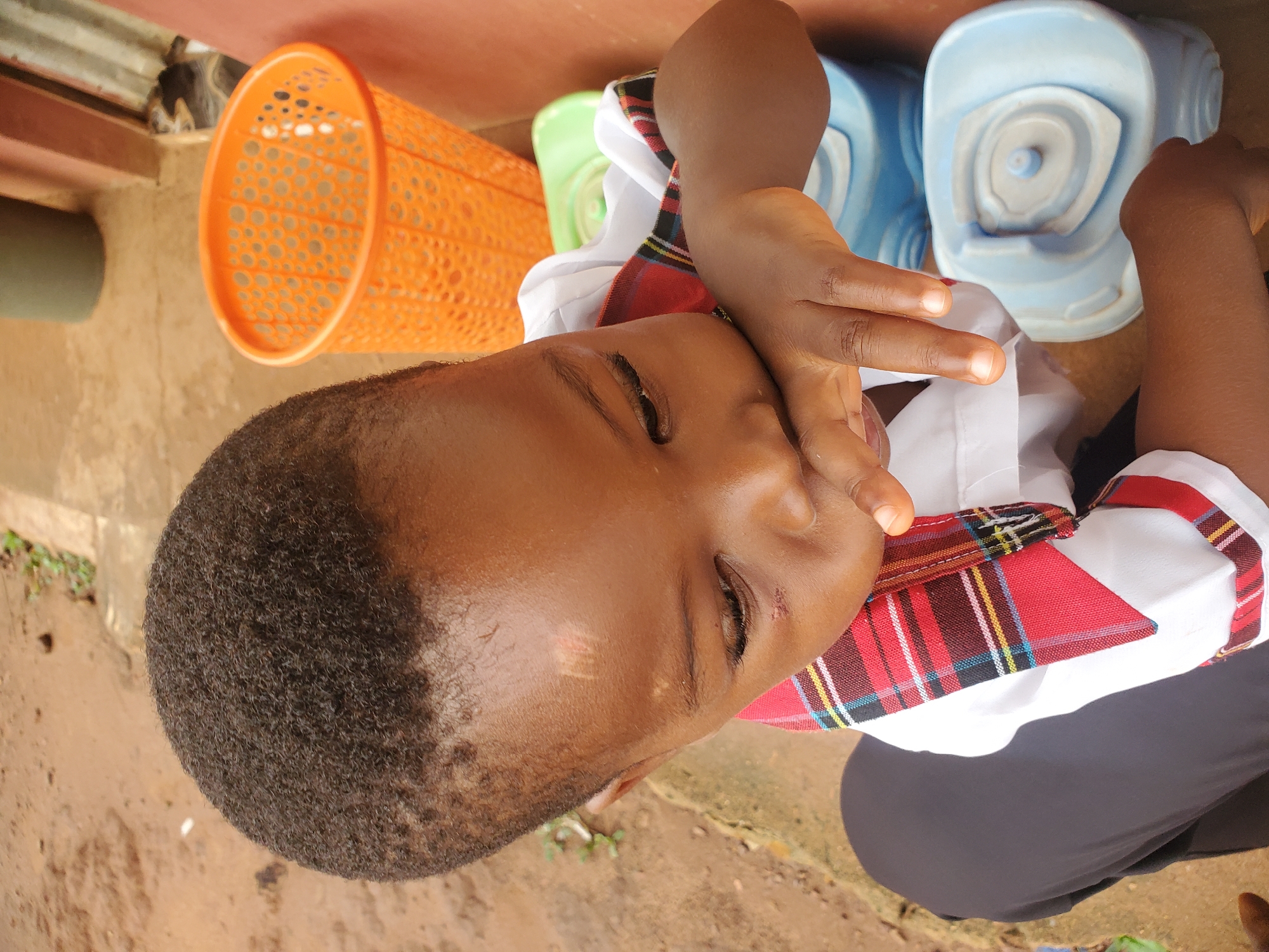 A young Black child in Ukwu-Abwa, Nigeria, rests their face on their hand. They are wearing a white shirt with a red, blue, and yellow plaid collar, over black pants. An orange plastic basket, green container, and blue toilet training seat sit nearby. The ground is visible, a mixture of dirt and concrete.