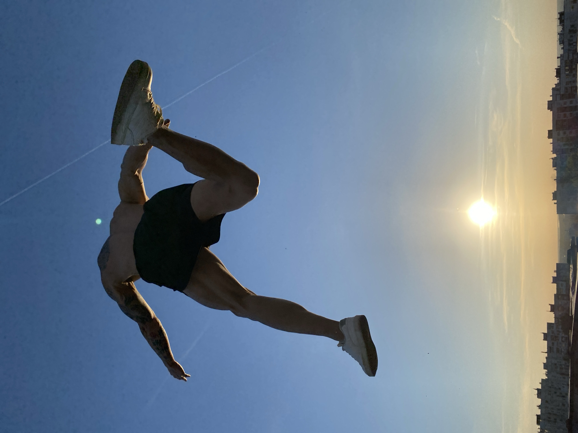A man is captured in mid-air against a bright sky, seemingly performing an acrobatic maneuver. He wears athletic shorts and shoes, with a tattoo visible on his left arm. The background features a cityscape silhouette under a sun-lit sky, likely during sunrise or sunset. The location is Pleven, Bulgaria.