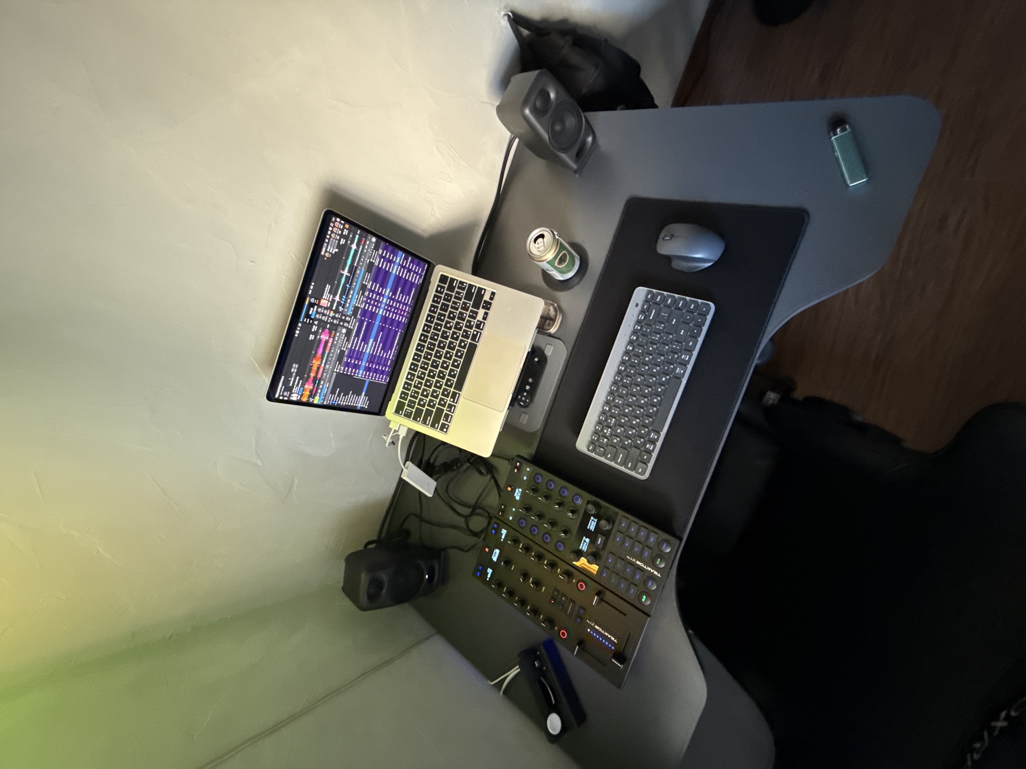 The image presents a workstation setup from an elevated, oblique angle.
The desk is gray, possibly metallic, and features an unusual, multi-tiered shape. On the upper tier, there is a small speaker, a can of alcoholic beverage, a mouse on a mouse pad, and a compact keyboard. On the lower tier, there is a laptop with a secondary monitor attached. The screen is displaying a complex visual interface. Further down on the lower tier is an audio mixing console, and near the bottom of the image, a black charging case.
The setting appears to be indoors, possibly a home office. Walls are pale, with a textured finish, perhaps plaster. The flooring is wood. The lighting is subdued, with varying levels of illumination.
There's no evidence from the image to suggest a location in Moscow, Russia.