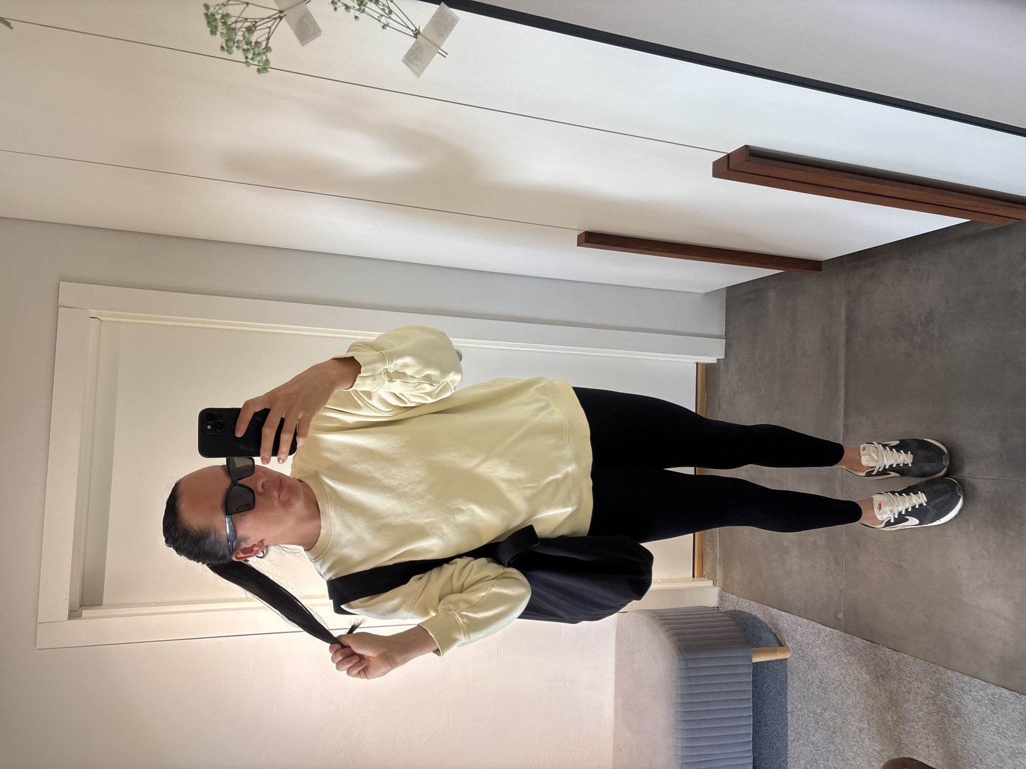 A person stands in front of a mirror taking a selfie. The person has their dark hair pulled back in a low ponytail and wears dark sunglasses. They wear a light yellow sweatshirt, black leggings, and black and gray sneakers. A black bag is slung over their shoulder. The environment appears to be a brightly lit interior with white walls and a gray floor. A gray upholstered bench is visible in the background. The scene is located in an unknown location. There is no evidence to suggest the location is in the city of Chaikovsky, Russia.