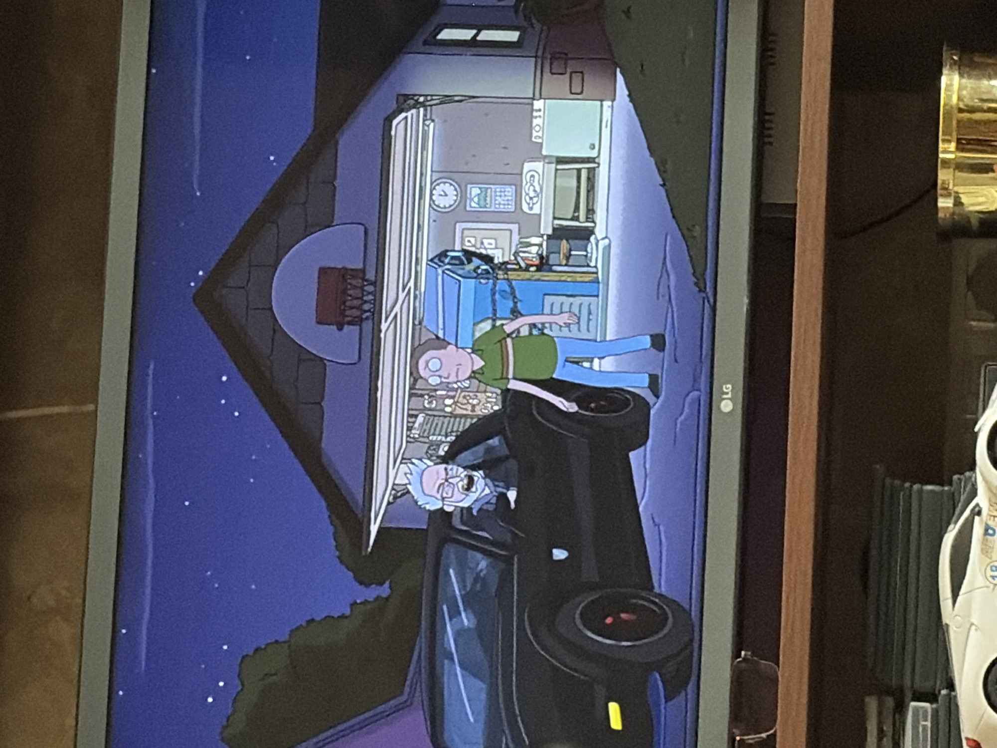 This is a screenshot from the animated television series Rick and Morty, depicting an outdoor scene at night in front of a suburban house. In the foreground, a black muscle car is parked, with Rick Sanchez in the driver's seat and Morty Smith standing beside the open car door. The car's headlights are on. Behind them, a garage door is open, revealing the interior of the garage which contains various tools and equipment, including a blue machine, a clock, and a calendar. A basketball hoop is visible above the garage door on the roof of the house. The sky is dark blue with visible stars. The scene is illuminated by ambient light and the car's headlights.
