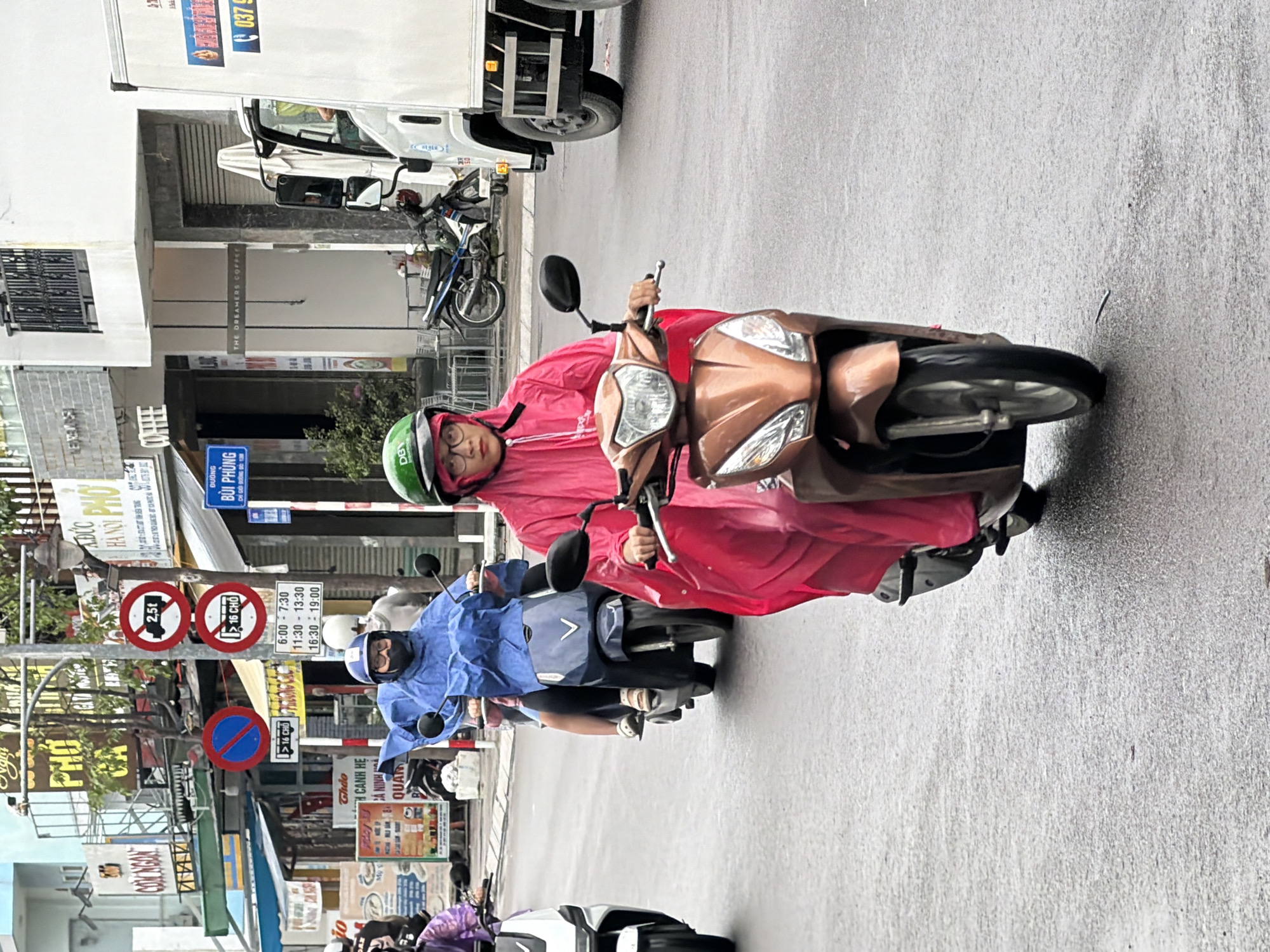 The image shows a street scene in Nha Trang, Vietnam, likely during a rainy day, as indicated by the wet road and the presence of people wearing raincoats. In the foreground, a person wearing a bright pink raincoat and a green helmet is riding a scooter. The scooter is a bronze color. Behind this individual, another person in a blue raincoat and a blue helmet is riding a dark-colored scooter.
The background reveals a street lined with shops and buildings, some with signs in Vietnamese. Several traffic signs are visible, including a circular red sign with a horizontal bar and a figure inside, likely indicating a weight restriction or a no-entry zone for certain vehicles. Another sign is circular with a red border and a blue center with a red diagonal line, which often signifies a prohibition. There are also signs indicating parking restrictions or times.
A white truck is parked or driving on the right side of the image. The general environment appears to be an urban setting with a moderate amount of commercial activity. The presence of multiple scooters suggests this is a common mode of transportation in the area. The overall atmosphere is muted, possibly due to overcast weather.