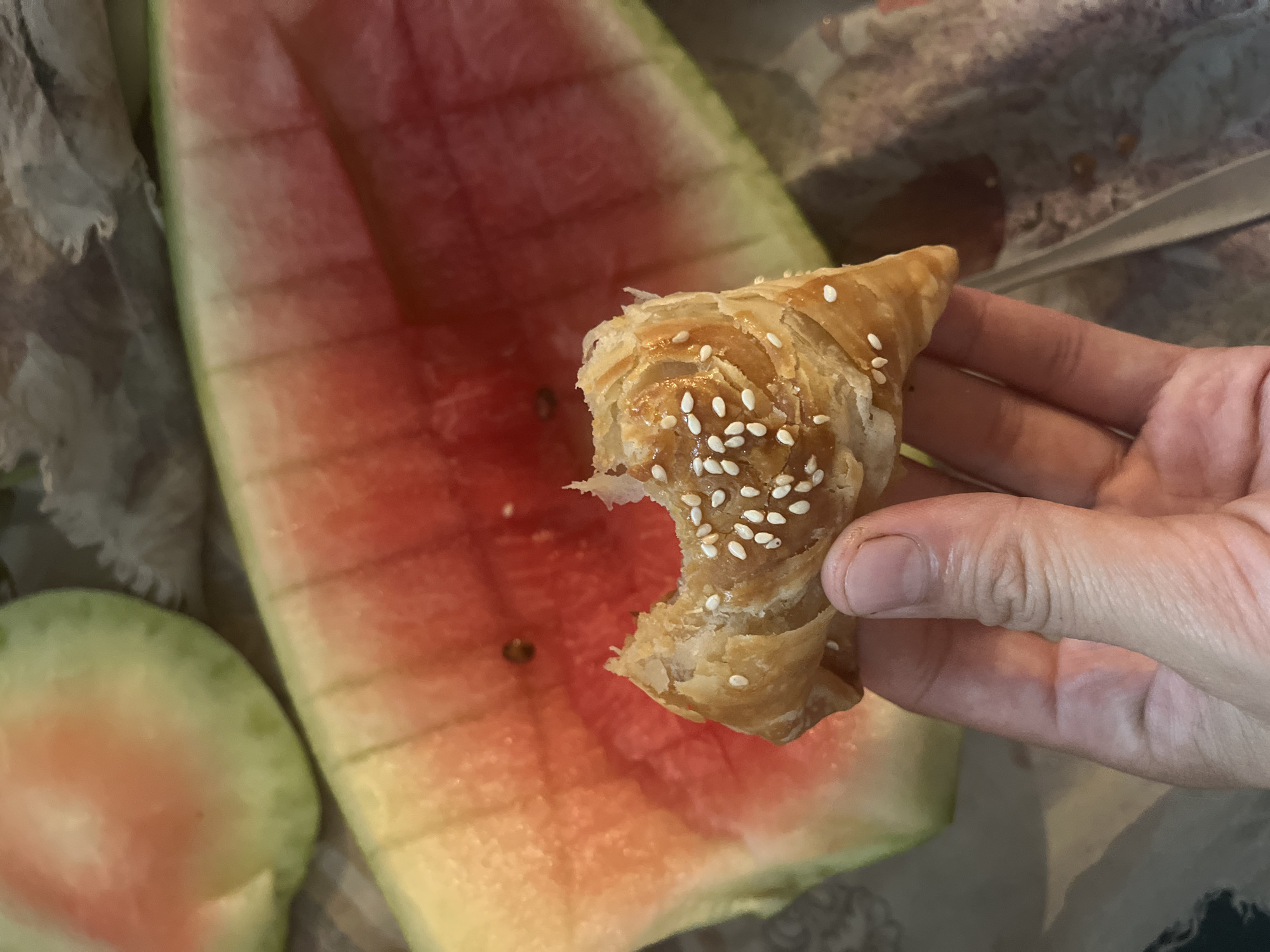 A hand holds a partially eaten pastry sprinkled with sesame seeds. In the background, a slice of watermelon is visible. The scene appears to be outdoors, possibly in a city like Tashlak, Kyrgyzstan, given the context provided. The lighting suggests daytime.