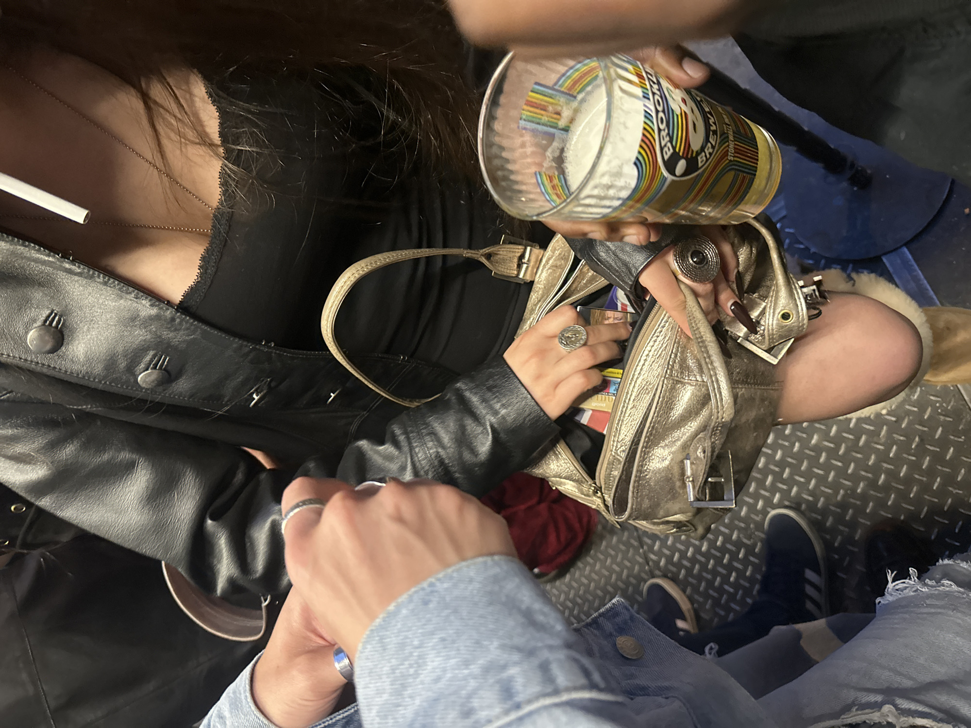 The photograph is a high-angle, close-up shot of a social gathering involving at least three individuals, possibly more. The individuals are partially visible and are engaged in activities related to socializing.
On the left, a person wearing a black leather jacket and a black top is holding a lit cigarette. A gold necklace is visible. This person's hand is touching the hand of another person, who is wearing a light blue denim jacket. This suggests an interaction between the two individuals.
A person with dark skin holds a glass of beer with a "Brooklyn Brewery" logo. A silver handbag sits on the lap of this person, who is wearing black tights or leggings.
The environment seems to be a crowded indoor setting, possibly a pub or a bar. The floor is made of a textured metal, and there is some seating visible in the background, including blue seats. Two pairs of shoes are visible; one is a pair of dark-colored sneakers, and the other is partially visible and is lighter in color.
Without additional context, pinpointing the exact location in London is impossible.