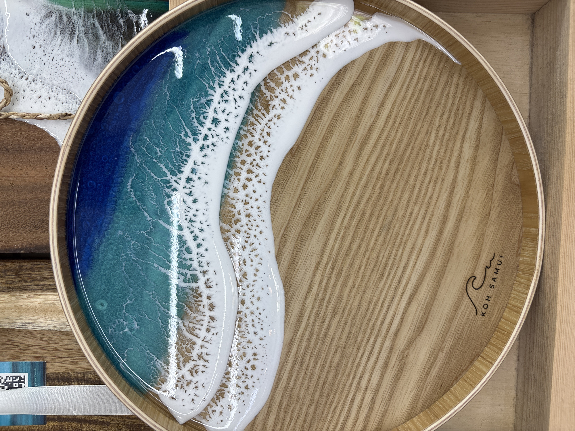 The image shows a wooden circular tray with a resin ocean art design. The resin is a gradient of deep blue to turquoise, representing the sea, followed by white resin simulating waves breaking on a beach. The remaining area of the tray is a light brown wood with visible grain. In the lower right quadrant, the words "KOH SAMUI" are inscribed in black next to a stylized wave. The tray is set within a wooden crate or box, with other wooden items visible on the sides. The location is indicated on the tray as Koh Samui, suggesting a connection to that location, which is near บ้านบางรักษ์, Thailand.