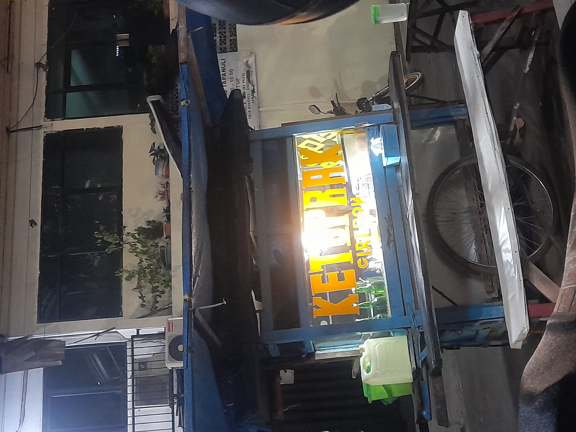 A street food cart vendor is pictured in the city of East Jakarta, Indonesia. The cart is blue and has a clear glass front displaying bottles of drinks and a lit sign that reads "KETOPRAK CILIBON." The cart is situated on a street at night, with a building visible in the background. A motorcycle is parked behind the cart. A white bench and a metal table are set up in front of the cart. The overall scene depicts a typical street food vendor setup in an urban environment.