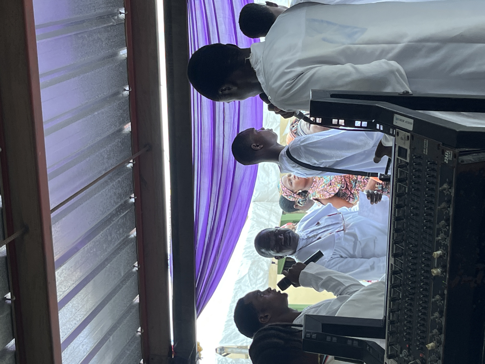 The image depicts an indoor scene with several individuals gathered, possibly for a religious or ceremonial event. The lighting suggests it is daytime. The setting appears to be a covered area with corrugated metal roofing visible on the left, suggesting a functional or open-air structure.
In the foreground, a person is holding a microphone and speaking into it, their face angled upwards. To their left and slightly behind, another person is also facing upwards, seemingly listening. Further back, more individuals are visible, some wearing white garments. A prominent purple curtain or fabric is draped behind the people, creating a backdrop. On the right side of the image, a piece of audio equipment, likely a mixing board, is visible with numerous knobs and buttons.
One person in the back is wearing a head covering with a colorful, patterned fabric. Another person, wearing white, has their hand near the mixing board, possibly operating it. The overall mood seems serious and focused, consistent with a formal gathering or service. The image is oriented vertically, and the people are also facing upwards as if looking at something or someone above them. There is a bright light source on the far left, which could be natural light or artificial lighting. The texture of the corrugated roof suggests a somewhat rustic or practical environment.