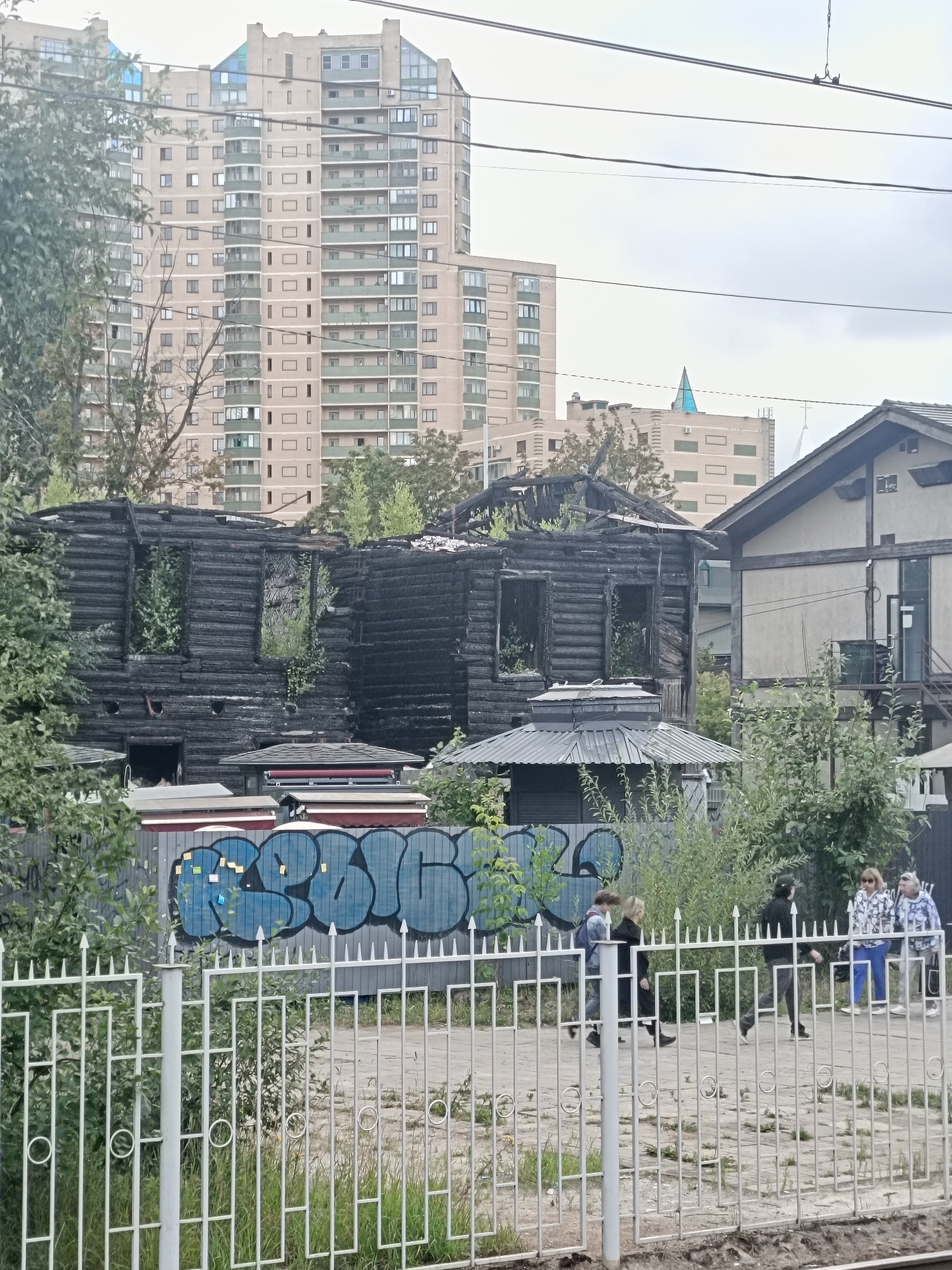 The image depicts a street scene in Saint Petersburg, Russia, with a burnt-out wooden building in the foreground, contrasted by a modern high-rise apartment building in the background. The wooden structure is severely damaged, with blackened walls and gaping holes where windows once were. Overgrown vegetation is visible emerging from the wreckage.
To the right of the burnt building, a partially intact, lighter-colored building stands. A white metal fence runs across the lower part of the image, separating the viewer from a sidewalk where four people are walking. The people are dressed in casual clothing. The sidewalk is paved, and there is a fence behind it with graffiti featuring large blue lettering. The overall environment suggests an urban setting, possibly undergoing redevelopment or experiencing neglect in parts. The sky is overcast, indicating a cloudy day.
