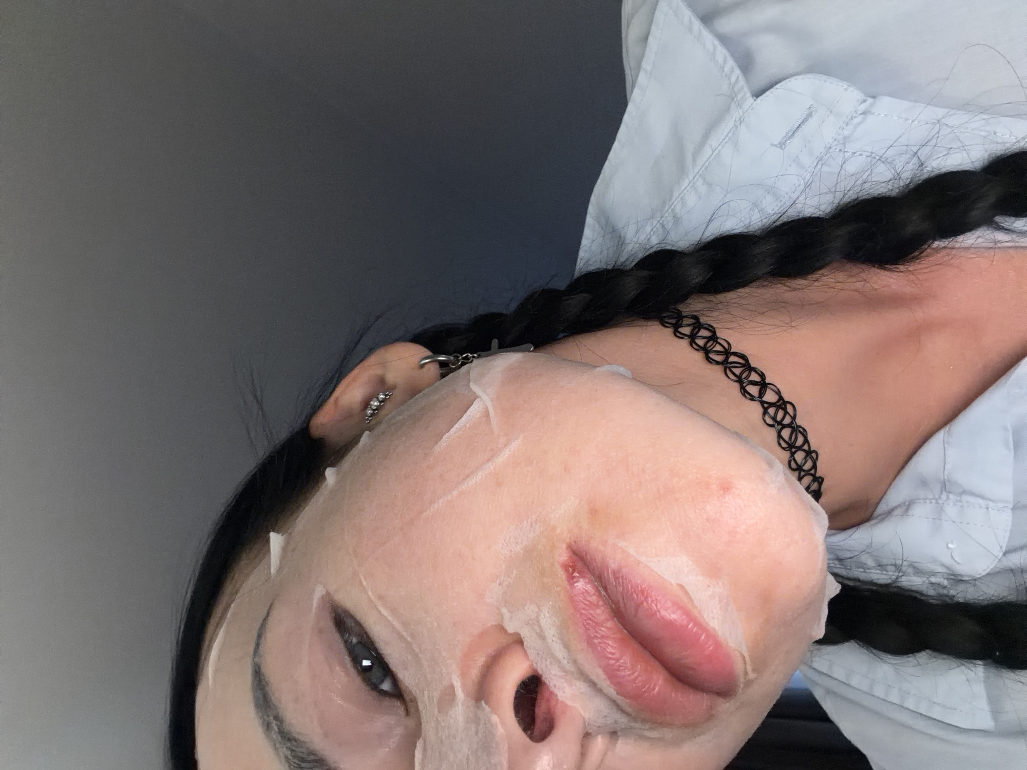 A close-up, low-angle shot shows the face of a young person wearing a white sheet mask. The mask covers most of the face, leaving the eyes, nose, and lips exposed. The person has dark hair styled in two braids, one of which falls over their shoulder. They are wearing a light blue collared shirt and a black choker necklace. The background is a plain gray wall. The lighting is dim, casting shadows on the person's face. The overall mood is intimate and relaxed.