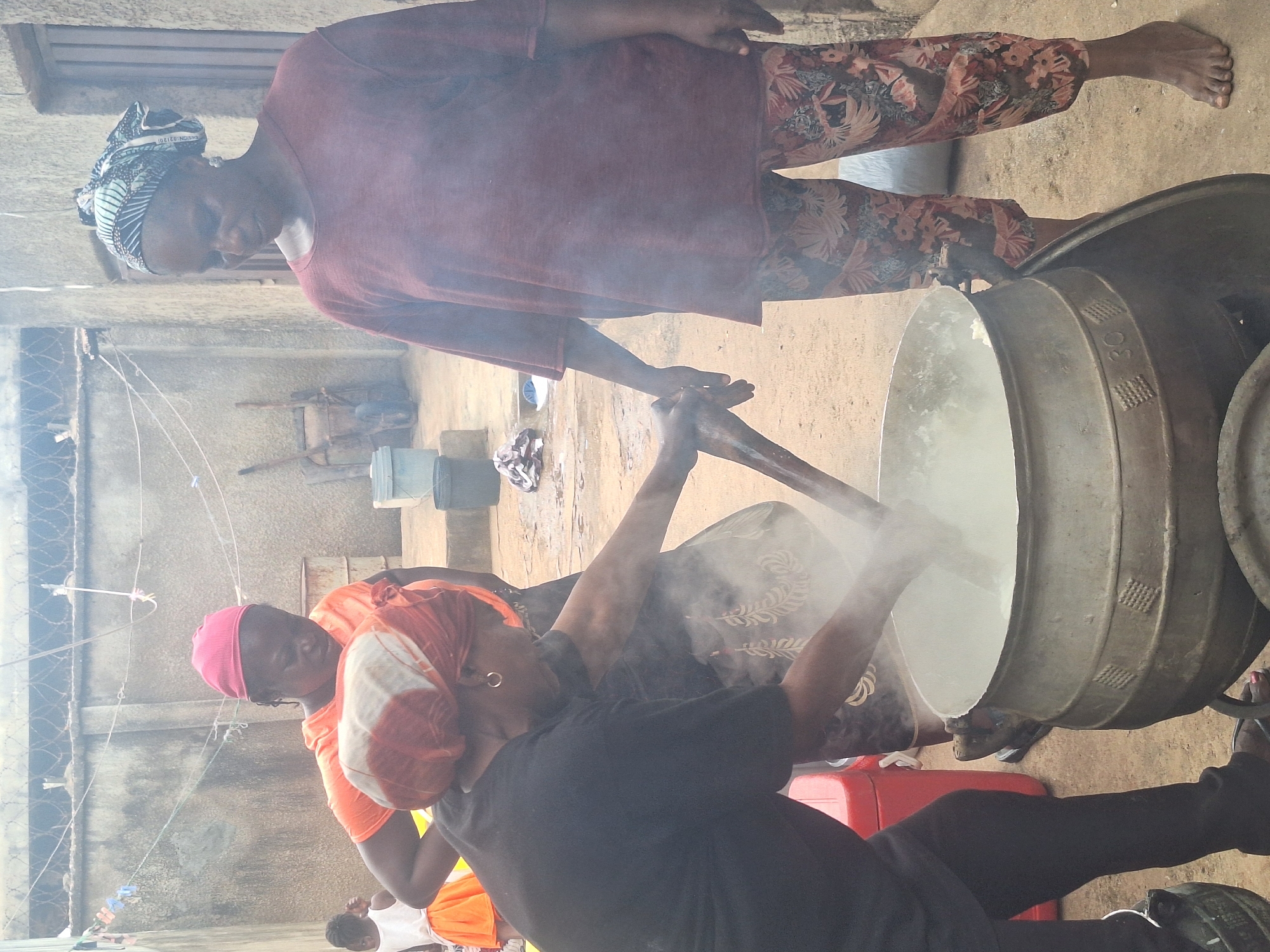 This image depicts a vibrant communal cooking scene in an outdoor or semi-outdoor setting in Sabon Tasha, Nigeria. At least three women are actively involved in preparing a large meal, utilizing traditional methods and large cooking vessels.
In the foreground, a woman wearing a black t-shirt, black trousers, and an orange and white patterned headwrap, with hoop earrings, vigorously stirs a large, metallic pot. Visible steam rises from the pot, indicating active cooking, likely boiling or simmering a substantial dish. She uses a long, sturdy wooden paddle for this task. To her left, partially obscured, is another woman in an orange top and a pink cap, observing or assisting with the cooking. The head of a small child is also visible near the bottom left of the frame.
Further back and slightly to the right, a third woman stands, observing the cooking process. She is dressed in a maroon long-sleeved top and distinctively patterned trousers featuring brown, red, and beige floral or leafy motifs. Her hair is covered by a patterned headwrap in black, white, and blue, and she wears earrings. Her feet are bare.
The central objects are two very large, industrial-style metallic pots or cauldrons, with one actively being stirred and another similar pot positioned next to it. The active pot displays the marking "3C" and textured grid-like patterns on its side. Red plastic jerry cans are visible on the ground nearby. The ground itself appears to be a mix of dirt and concrete.
The background reveals elements of a residential or communal area, including walls, what appears to be a fence with barbed wire on top, and clotheslines strung with pegs between structures. The lighting suggests it is daytime, with diffused light indicating either an overcast sky or a shaded area. The overall atmosphere conveys a sense of focused activity and shared effort in preparing food, highlighting community and traditional practices. On the headwrap of the woman in maroon, the text "031201" is faintly visible.