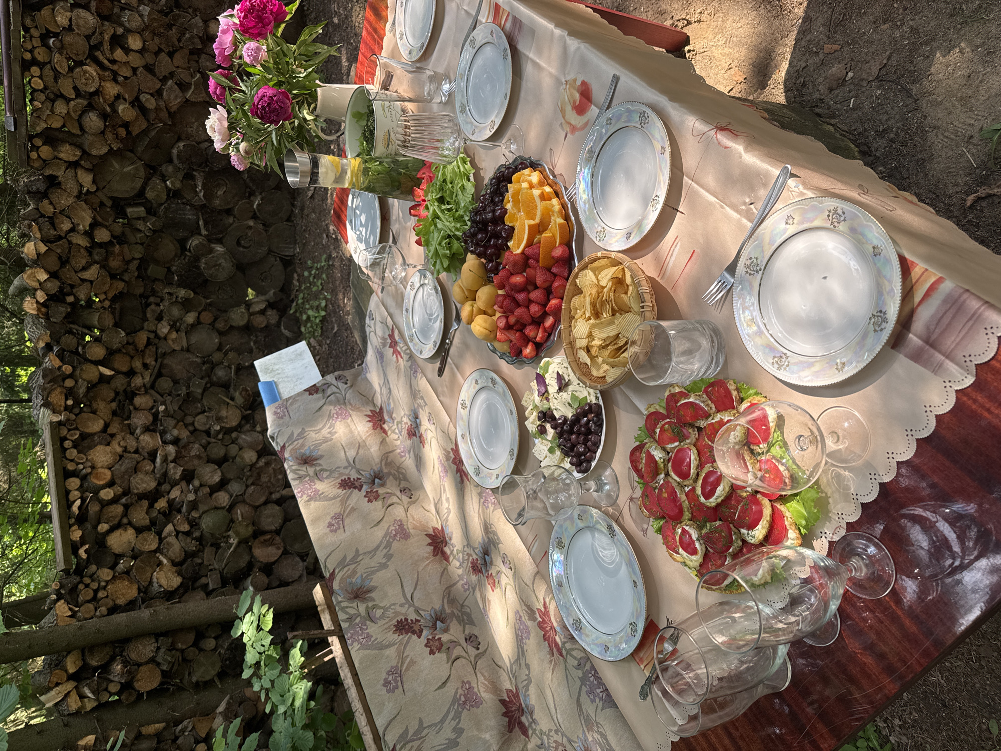 An outdoor table set for a meal is photographed from a slightly elevated angle. The table is covered with a tablecloth patterned with floral motifs. On the table are several plates, glassware, and various food items including a platter of fruit, a dish of cheese and olives, a bowl of chips, and a small plate of canapés. In the background, a stack of firewood is visible, and beyond that, trees suggest a forested environment. The location is identified as Козино, Russia. There are no people present in the image.