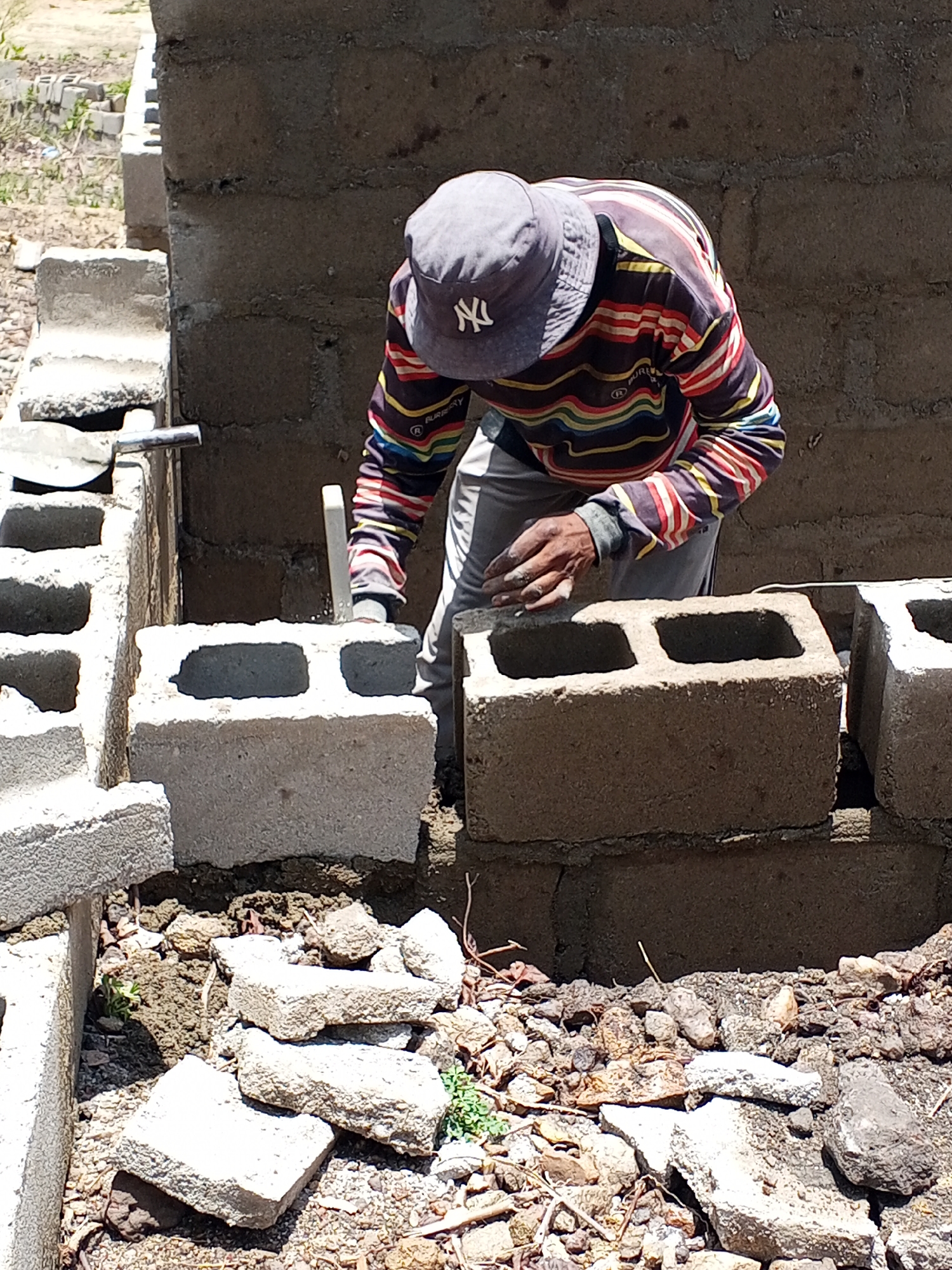 A man is laying cinder blocks to build a wall in Lokoja, Nigeria. The man is wearing a striped long-sleeved shirt, grey pants, and a grey baseball cap. He is bending over and using his hands to position a cinder block on top of a partially built wall. There are other cinder blocks scattered around him on the ground, along with pieces of stone. A trowel is seen inserted in-between 2 blocks. The partially-constructed wall is made of similar cinder blocks. The background is blurred, showing foliage and other construction materials.