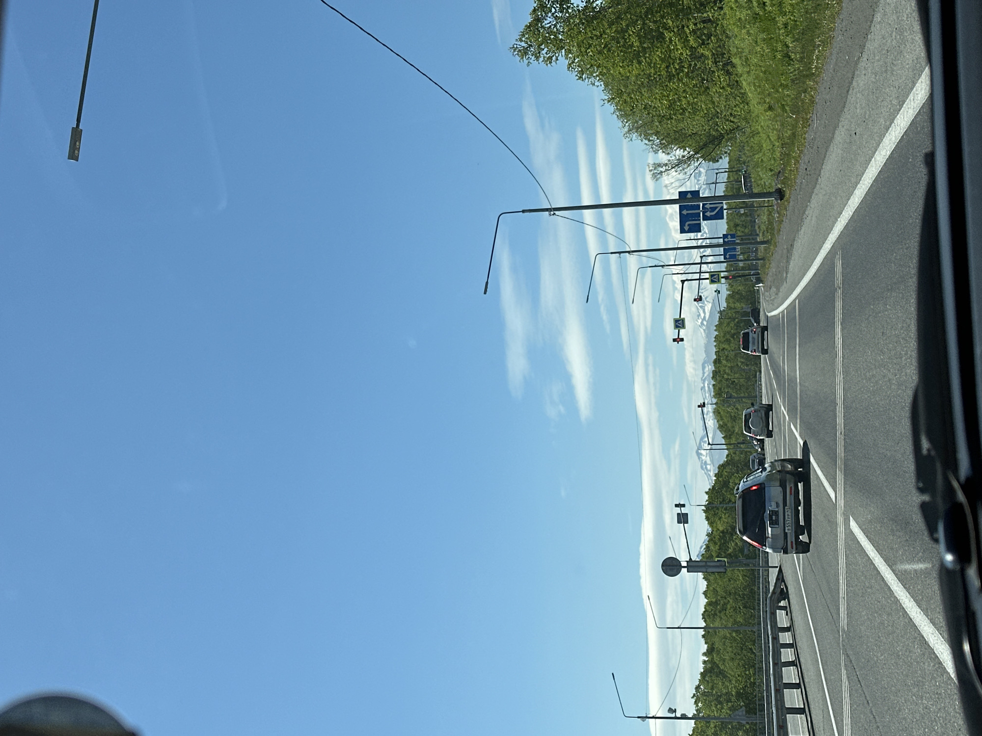 The image depicts a view from a vehicle traveling on a multi-lane road in the city of Svetly, Russia. The sky is clear blue with scattered white clouds. Several streetlights are visible along the roadside. On the right side of the road, lush green trees and vegetation line the embankment, with distant snow-capped mountains visible on the horizon. Traffic signs indicating lane directions are posted. Multiple vehicles are driving on the road, including a silver car in the foreground with a license plate that appears to read "B 557 PP 41". The road itself is asphalt with clear white lane markings. A metal guardrail is present on the left side of the road.