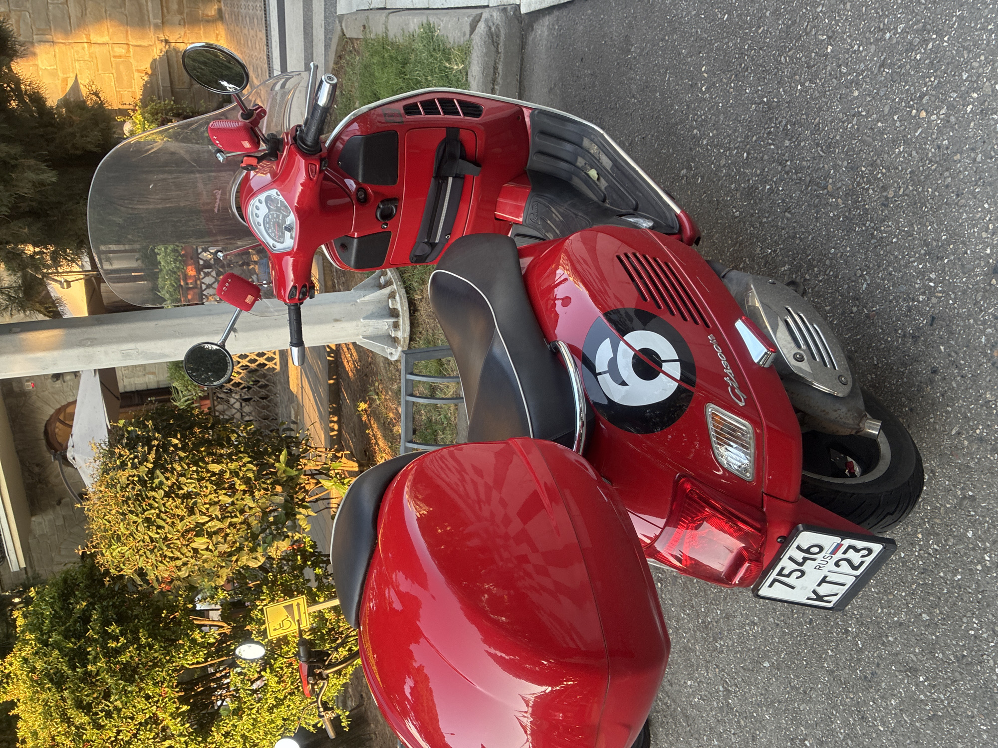 The image shows a red Vespa scooter parked on an asphalt surface. The scooter has a large red top box and a clear windshield. There is a circular black and white decal with the number "6" on the side of the scooter. A license plate is attached to the rear of the scooter, displaying "7546 RUS KT 23". In the background, there are green bushes, a stone building, and a light pole. The scene appears to be outdoors, possibly in a public area. The lighting suggests it is daytime, with sunlight casting shadows. The scooter is not in motion. There are no people visible in the image.
