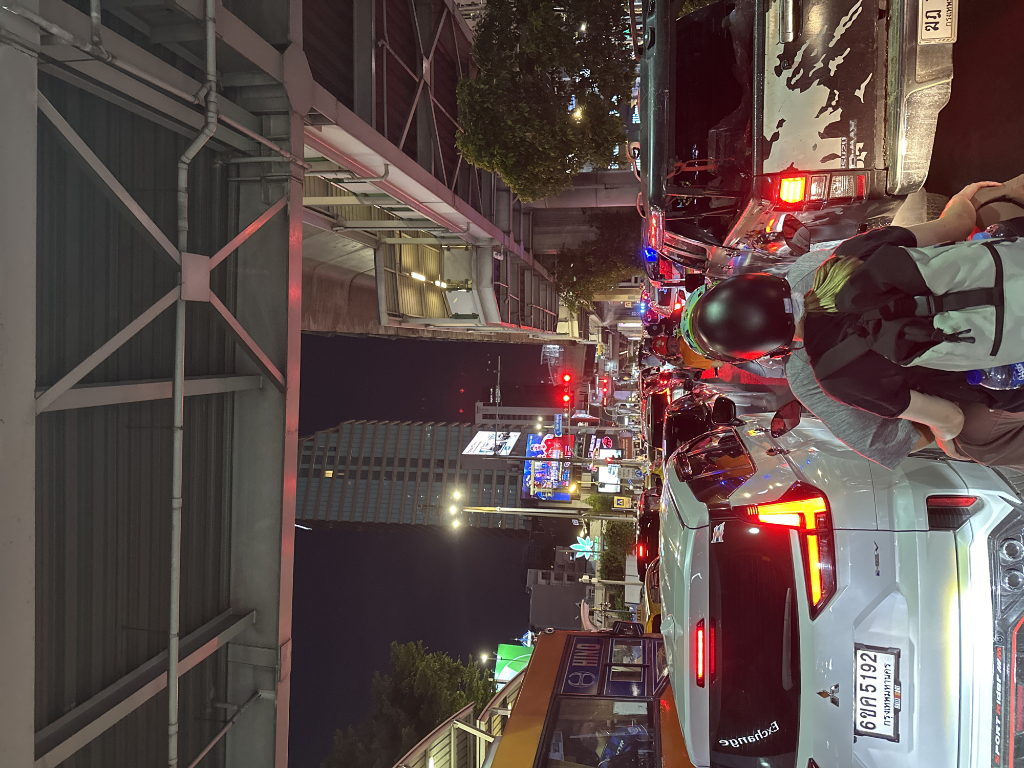 The image depicts a congested street scene in Bangkok, Thailand, at night. Heavy traffic is evident, with cars, motorcycles, and a bus tightly packed together. The street is lined with billboards, and the illuminated cityscape, featuring tall buildings, is visible in the background. A pedestrian overpass is prominent on the left. On a motorcycle in the foreground, a person wearing a helmet and carrying a backpack is waiting in traffic. Car license plates, traffic lights, and street signs are visible.