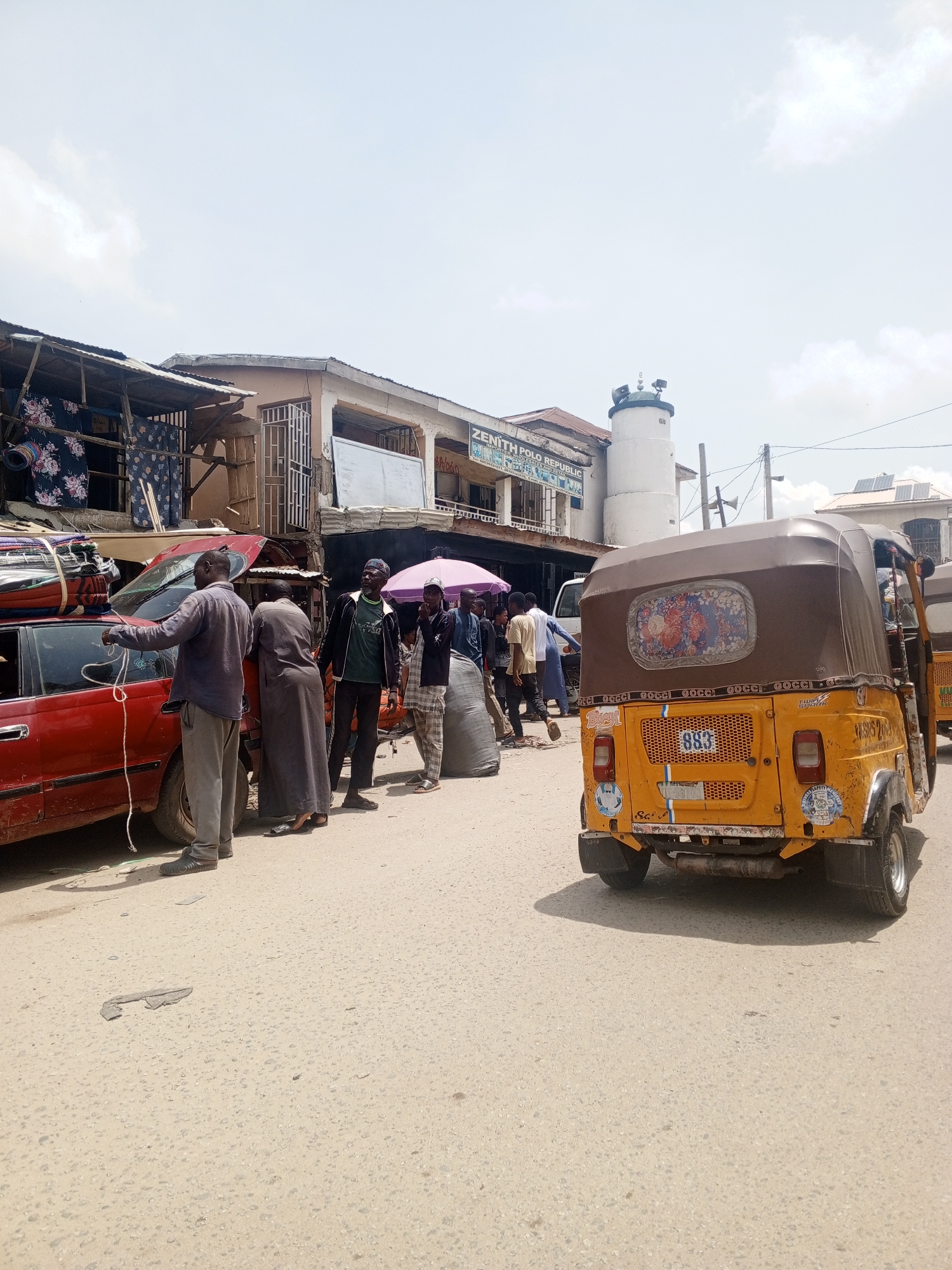 The image depicts a street scene in an urban area of Unknown, Unknown. In the foreground, a yellow auto-rickshaw faces away from the viewer. To the left, a red car is loaded with textiles. Several people are gathered around the car, including a man tying down the load with a rope, and others standing nearby. Further back, more people are seen along the street, some with an umbrella. The buildings along the street include businesses with signage, such as Zenith Polo Republic, and a cylindrical tower-like structure. Power lines are visible in the sky. The road is paved and appears to be heavily used.
