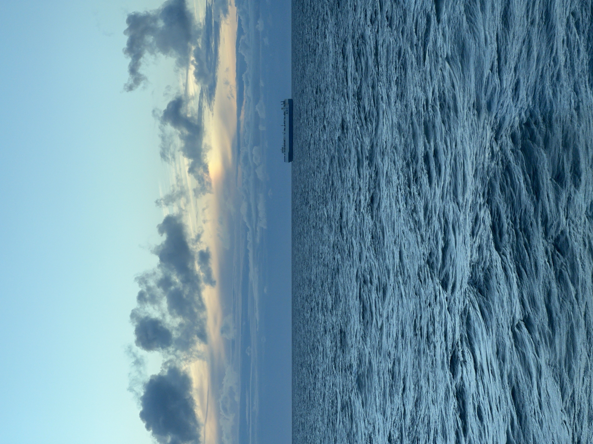A large cargo ship, identified by the letters "ARC" on its side, sails on a vast, dark blue ocean under a twilight sky. The sky is a gradient of pale blue at the top, transitioning to darker blues and grays with scattered clouds near the horizon. The water is choppy with visible waves, reflecting the muted colors of the sky. The ship is positioned in the middle distance, centered horizontally, and appears to be traveling from left to right. The scene is devoid of any other vessels or land features. The overall impression is one of solitude and the immensity of the sea. The location is Unknown, Poland.