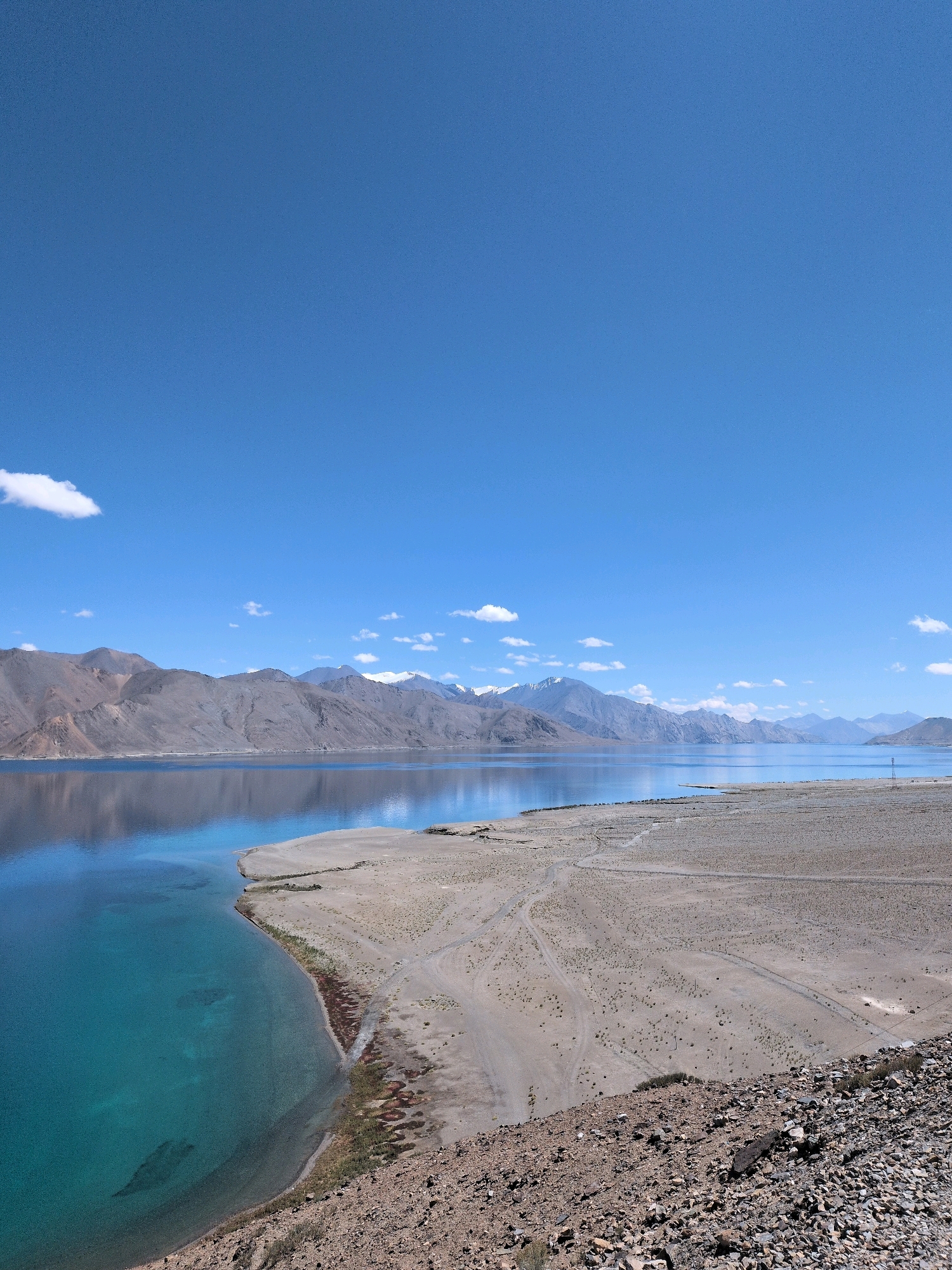 This is a high-angle, full shot of Pangong Tso lake in Ladakh, India, under a clear blue sky with scattered white clouds. The lake's water transitions from a vibrant turquoise near the shore to a deep blue further out, reflecting the surrounding mountains. The mountainous landscape on the far side of the lake is rugged and barren, with some snow-capped peaks visible in the distance. The foreground consists of a dry, rocky, and sandy terrain, with sparse vegetation. A thin, sandy strip of land juts out into the lake on the left side of the frame. The scene conveys a sense of vastness and natural beauty.