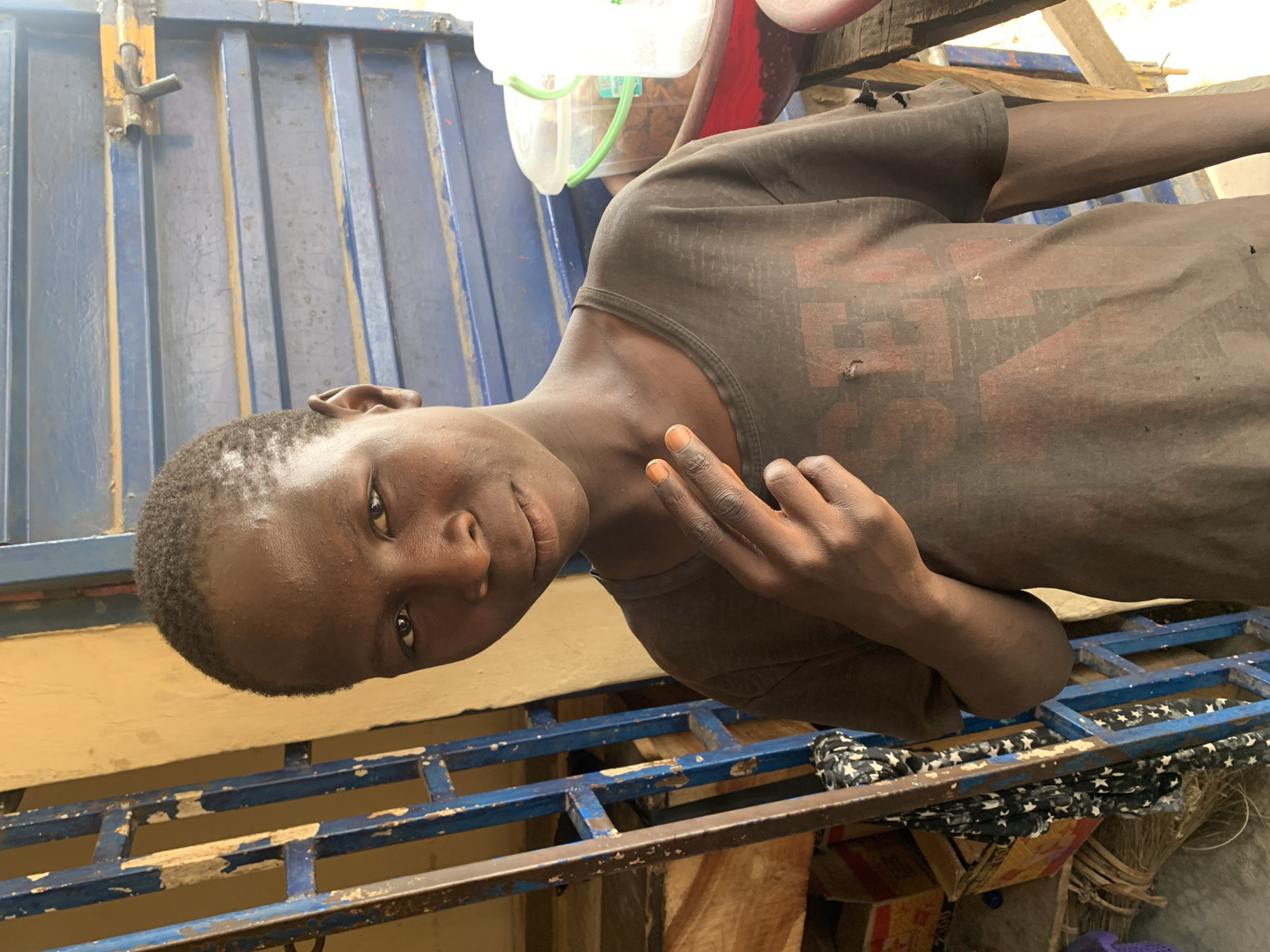 A young, dark-skinned person is seen in Jos, Nigeria, wearing a dark t-shirt with a faint red graphic. The background features a blue metal structure and a yellow wall. The person is holding their fingers up in a gesture. Other objects include a white container, a red object, wood, and a black and white star-patterned cloth. The environment appears to be an outdoor space.
