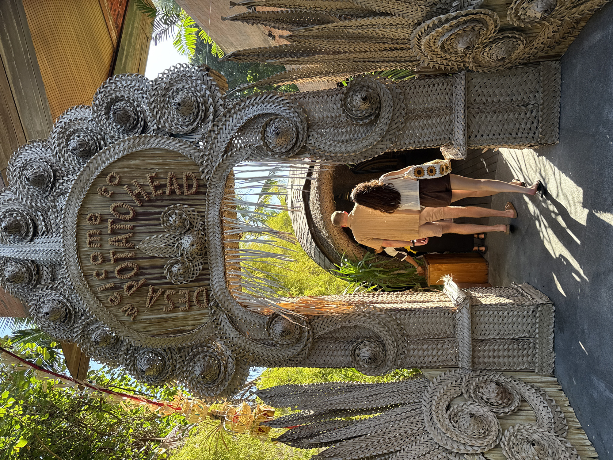 In North Kuta, Indonesia, an ornate archway woven from dried palm fronds serves as an entrance to a botanical garden. The arch is decorated with intricate spiral patterns and a circular sign made of woven wood and small, light-colored seeds. The sign reads "Welcome to BOTANICA". Two individuals are walking through the archway into the garden. The person in front is a man wearing a tan short-sleeved shirt and light brown shorts. Behind him, a woman is walking, wearing a white crochet tank top and dark brown shorts, carrying a tote bag adorned with a sunflower pattern. The ground is paved with dark grey material, and the sunlight casts long shadows from the archway and surrounding foliage. Lush green trees and foliage are visible beyond the arch, with a hint of brick architecture on the right side of the frame. The overall atmosphere is tropical and inviting.