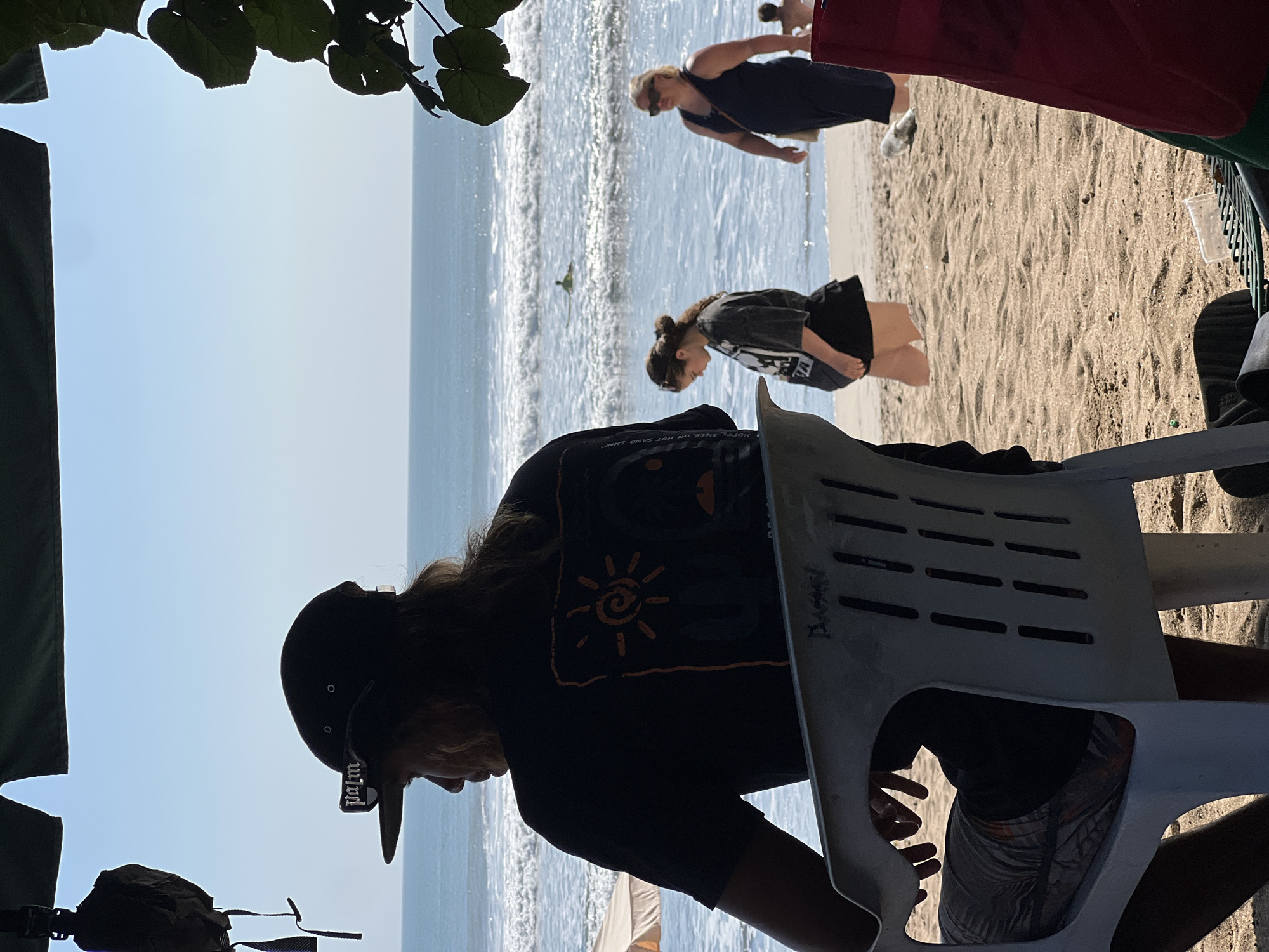 A man with long hair, wearing a black baseball cap with sunglasses perched on the brim and a black t-shirt with a sun graphic, sits in a white plastic chair on a sandy beach in Kuta, Indonesia. He is facing away from the camera, looking out at the ocean. Behind him, a woman in a dark t-shirt and black skirt walks along the beach, and further back, a woman in a dark blue dress walks towards the right. The ocean has gentle waves, and a surfer can be seen in the distance. Leaves from a tree hang down on the right side of the frame. The lighting suggests it is daytime, with clear blue skies.