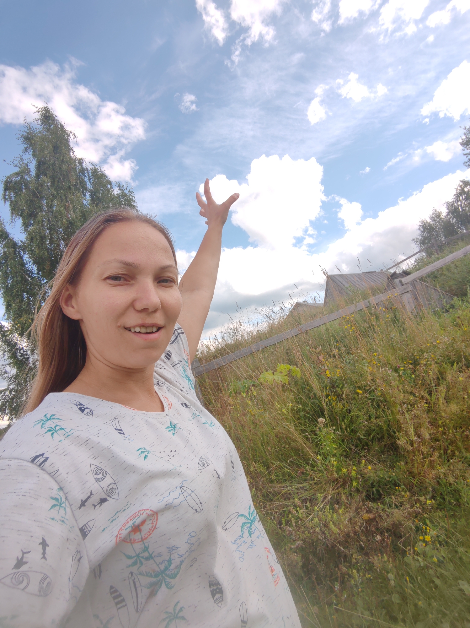 A woman with blonde hair is pictured in a close-up selfie outdoors. She is wearing a white t-shirt with a tropical print of palm trees, surfboards, and sharks. Her right arm is raised with her fingers extended towards a cloud in the blue sky. The background features a tree on the left, tall grass, and a wooden fence with some rustic structures behind it. The setting appears to be rural, consistent with the outskirts of Чухлэм, Russia. The lighting suggests it is daytime, possibly late morning or early afternoon.