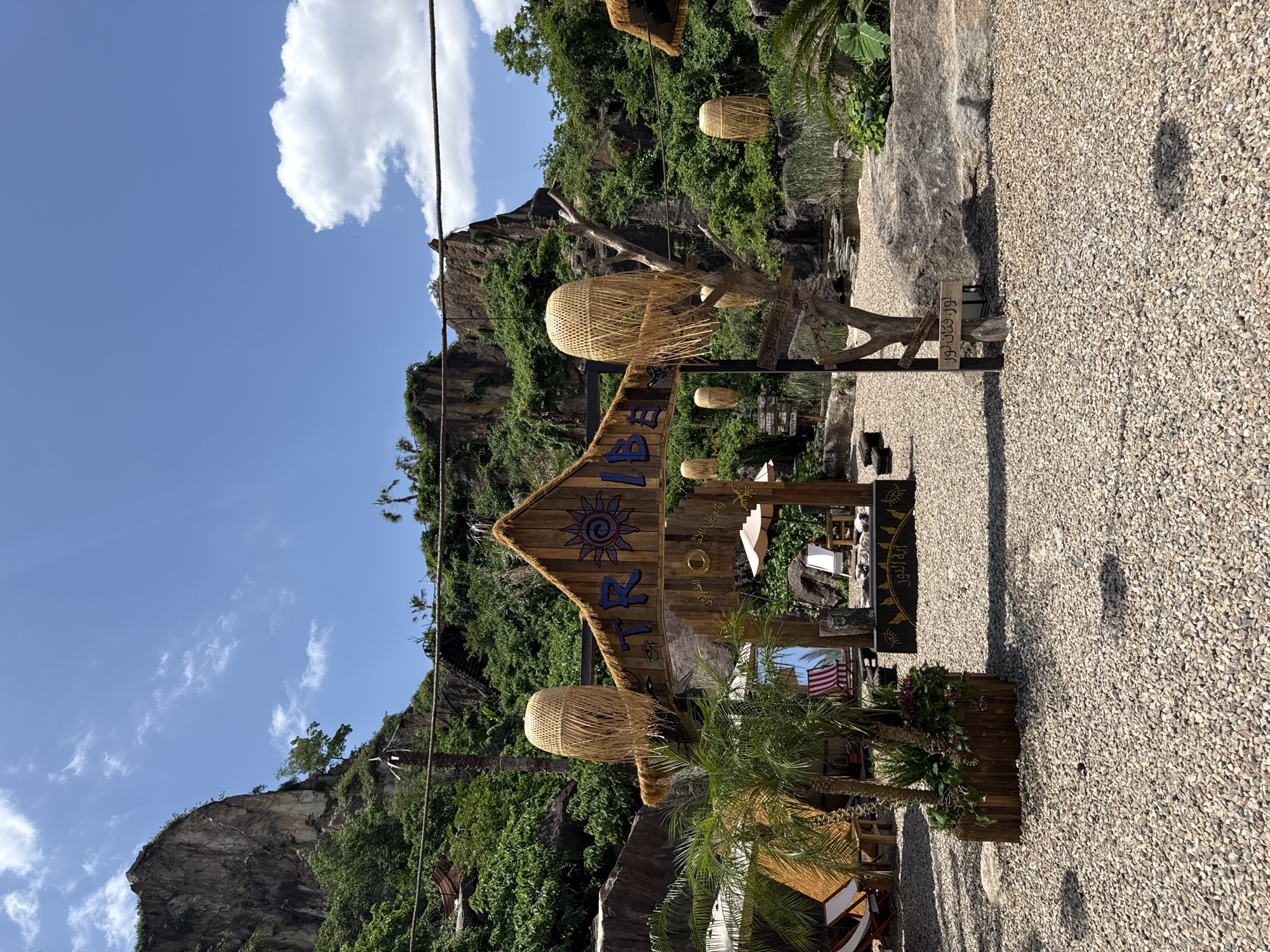 The image shows a scene in Keqiao District, China. The foreground features a wooden structure resembling an entrance sign with thatched roofing and hanging wicker lamps. The sign displays the name "TRIP" in blue lettering, accompanied by a sun symbol and other decorative elements. A planter box with a small palm tree and other greenery sits near the base of the sign. The ground is covered with small gravel.
In the background, a rocky cliff covered with lush vegetation rises into the sky. The sky is blue with scattered clouds. Cables run along the cliff face. Other wooden structures and decorative elements are visible further into the scene, suggesting a tourist or recreational area.