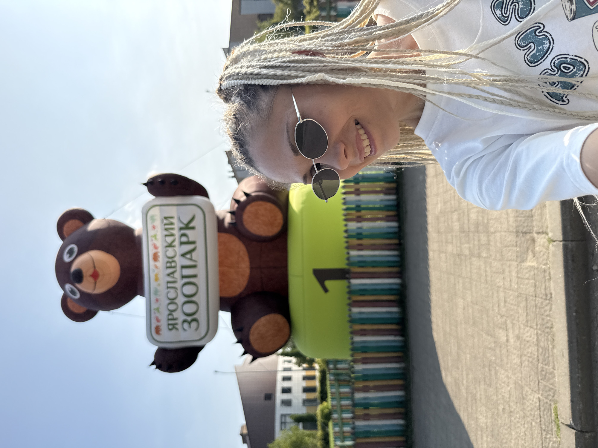 A medium shot shows a woman with blonde braided hair and sunglasses smiling in the foreground, with a large inflatable bear holding a sign that reads "ЯРОСЛАВСКИЙ ЗООПАРК" (Yaroslavl Zoo) in the background. The bear is perched on a bright green base with the number "1" visible. A colorful striped fence separates the paved area in the foreground from a grassy area with trees and buildings in the distance. The scene appears to be outdoors on a sunny day, likely at the Yaroslavl Zoo in Ignatovo, Russia.