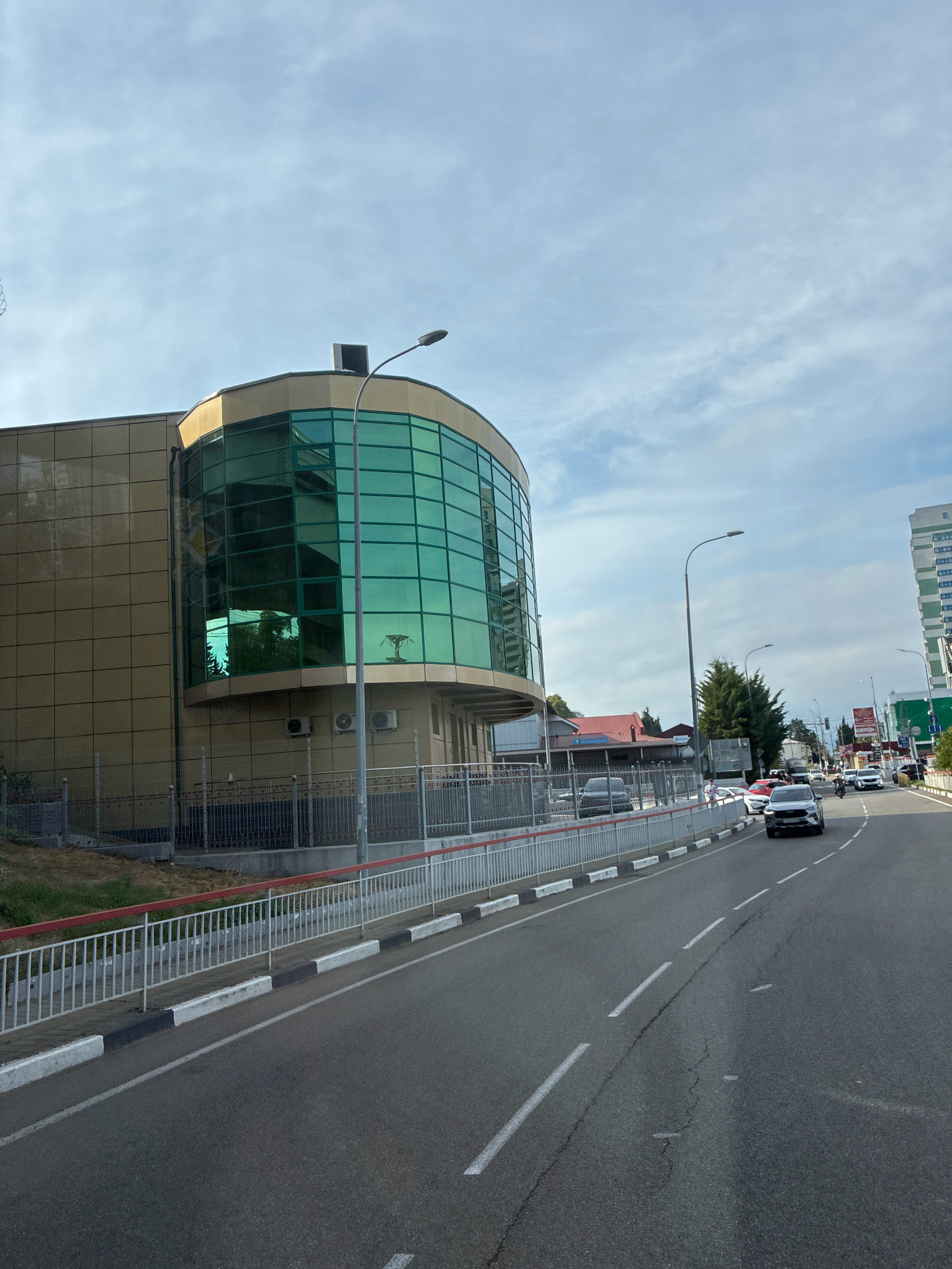 The image captures a street view in Sochi, Russia, featuring a modern building with a distinctive curved facade of green glass. The building is a light tan color with a tiled exterior. Adjacent to the building is a street with several cars driving on it, including a white SUV in the foreground. The street is bordered by a sidewalk and a metal fence with a red and white railing. In the background, more buildings and trees are visible under a cloudy sky. The overall scene suggests an urban environment on a bright, partly cloudy day.