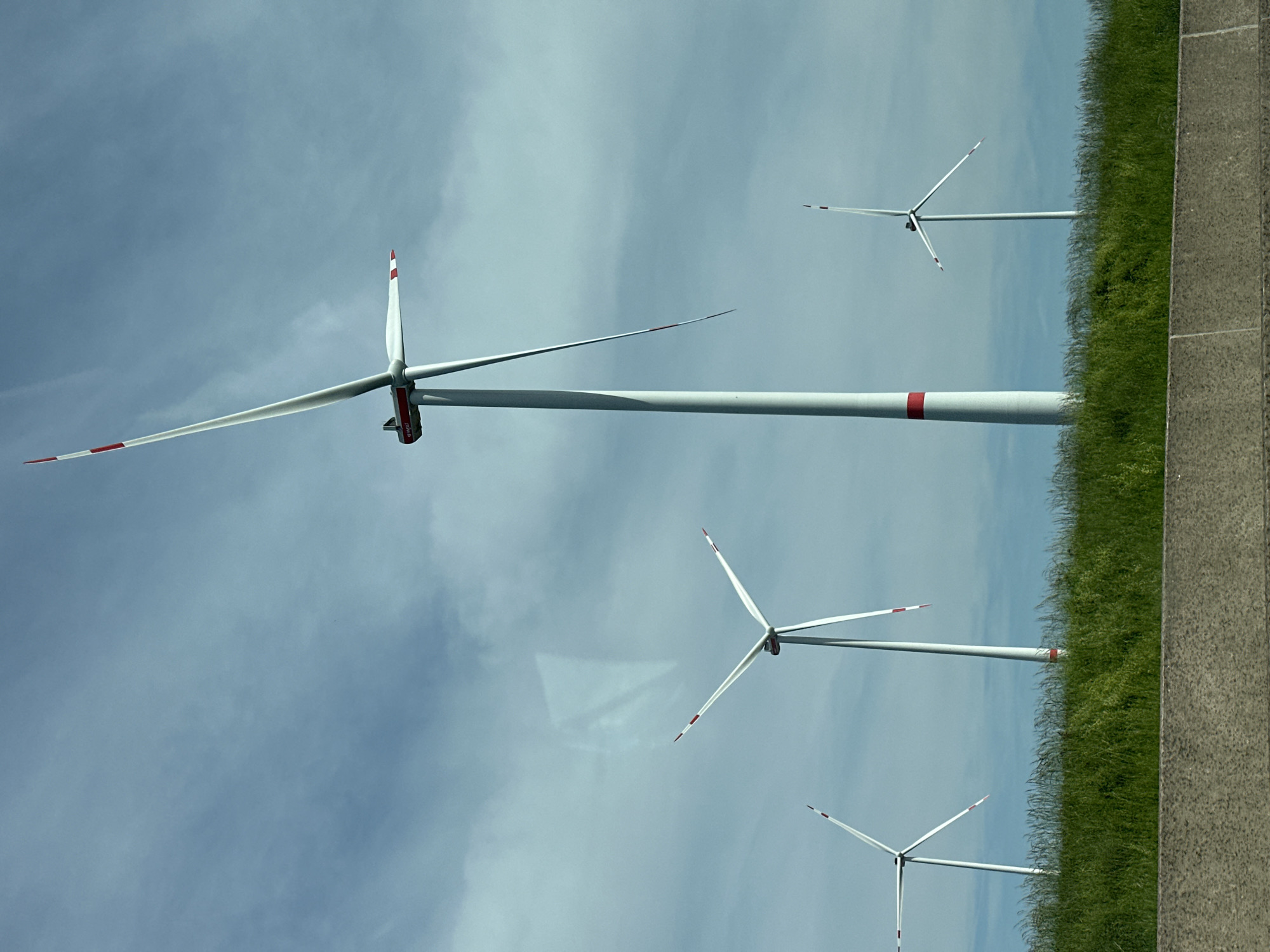 The image shows a view of wind turbines in Bockenem, Germany. The turbines are white with red markings on the blade tips and nacelles. There are four turbines visible, varying in size due to perspective. The foreground shows grass and a concrete surface. The background is a cloudy sky.