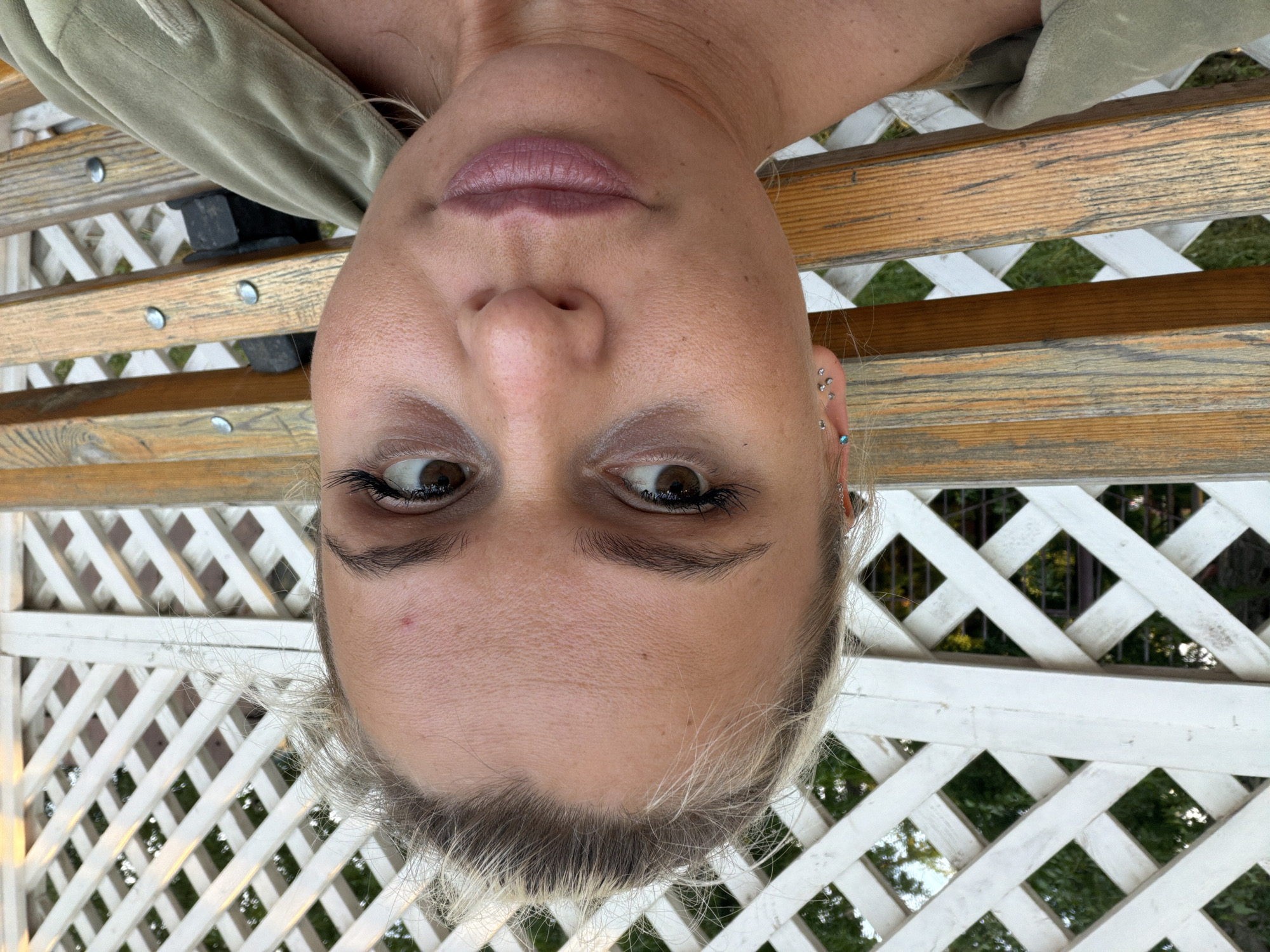 A close-up, upside-down portrait of a fair-skinned person with brown eyes and light blonde hair is set against a backdrop of a partially visible, light-colored wooden bench and a white lattice fence. The person has multiple piercings on their ear. The background shows glimpses of greenery through the fence. The location is presumed to be Sarapul, Russia, as per the prompt.
