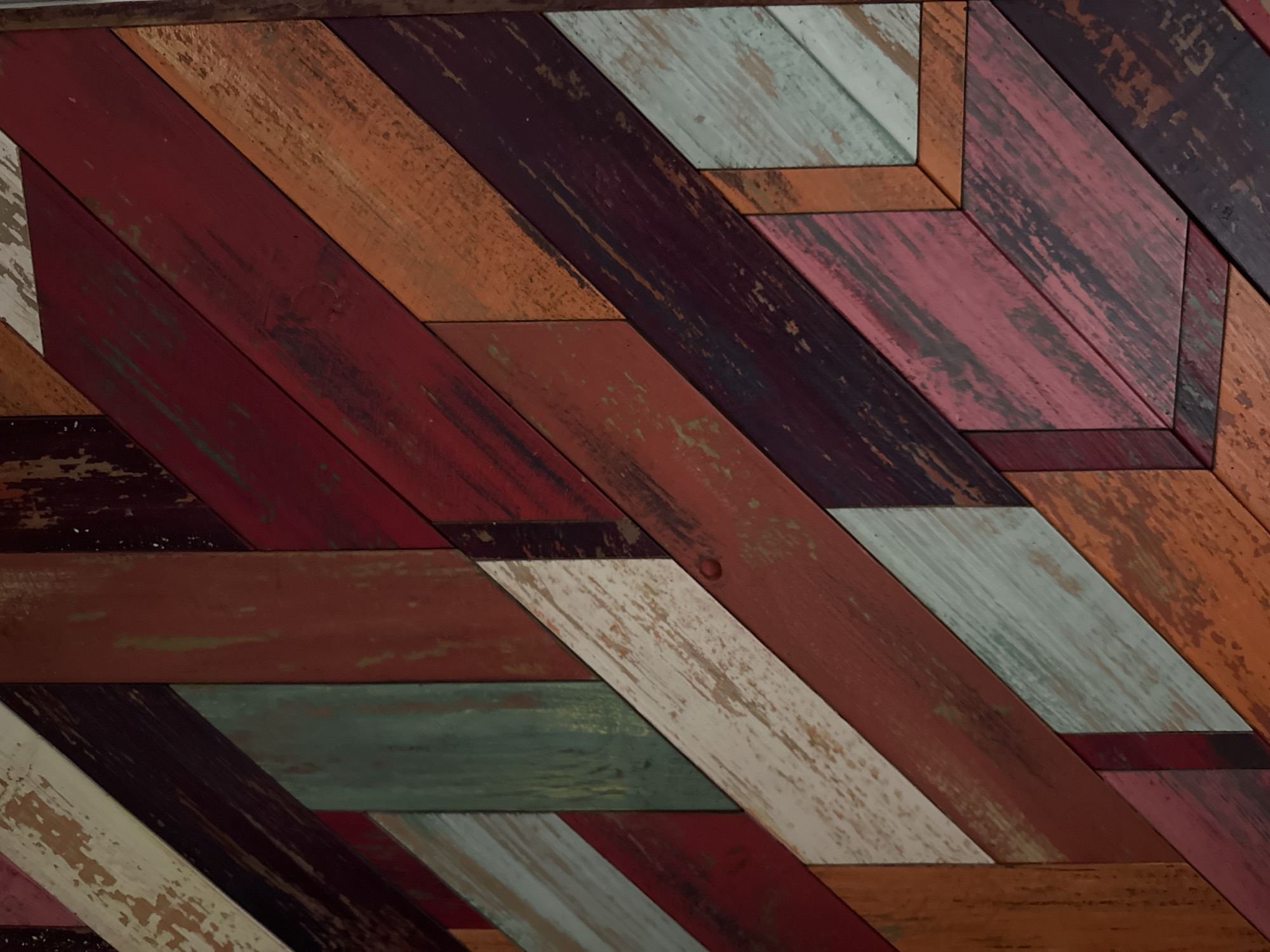 The image depicts a wall constructed of wooden planks of varying colors arranged in a diagonal pattern. The colors include shades of red, brown, orange, white, blue, and green. The planks have a distressed look, with some paint missing or peeling off. There are no people or other identifiable objects in the image. There is no information in the image to suggest this is located in Saint Petersburg, Russia.