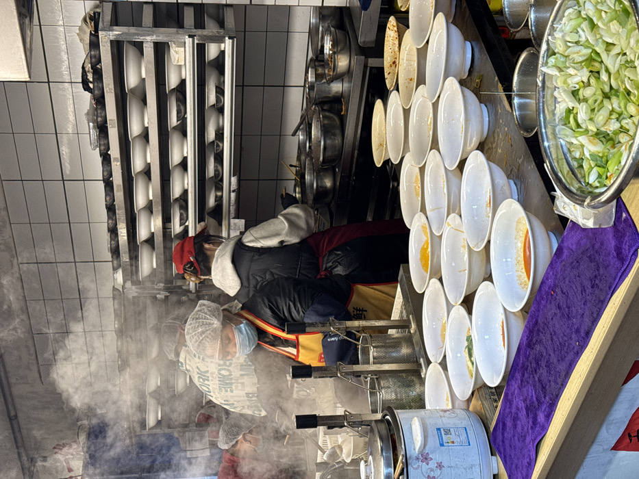 Inside a bustling commercial kitchen, cooks prepare food amidst rising steam. One worker wearing a hairnet and mask tends to a cooking apparatus. Rows of bowls line the counters, some containing colorful ingredients. A metal rack stores additional bowls. The kitchen's walls are tiled, and stainless steel equipment lines the work area. A bowl of chopped green onions sits atop a purple cloth.